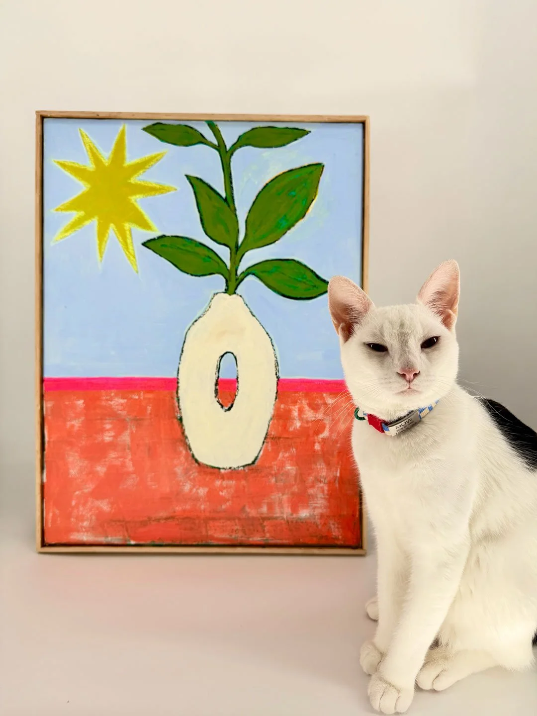 A white cat with a collar sitting next to a colorful abstract painting of a plant in a vase with a yellow star and a red surface background.