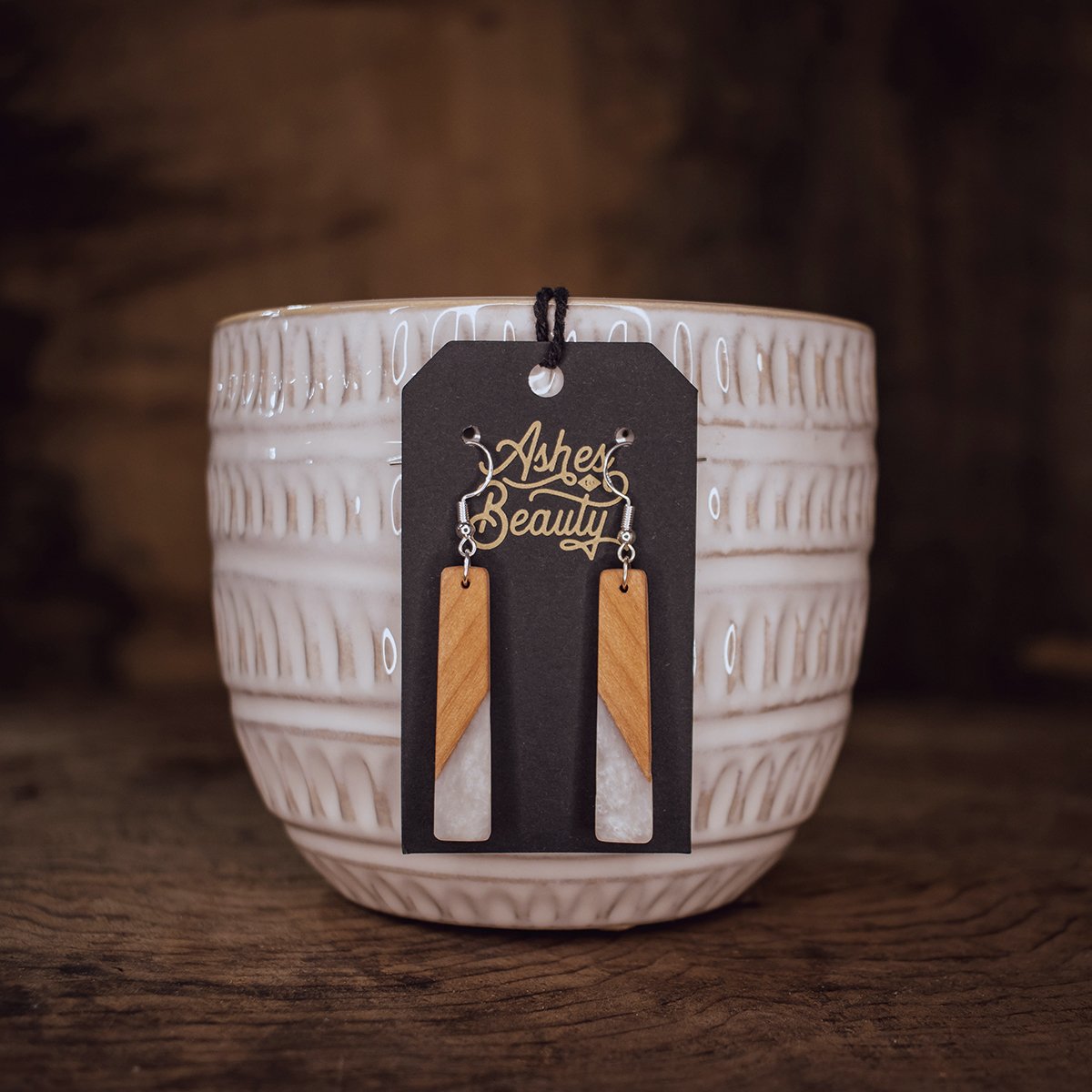 Earrings with wooden and marble elements displayed on a black card labeled 'Asher Beauty', held by a ceramic holder on a wooden surface.