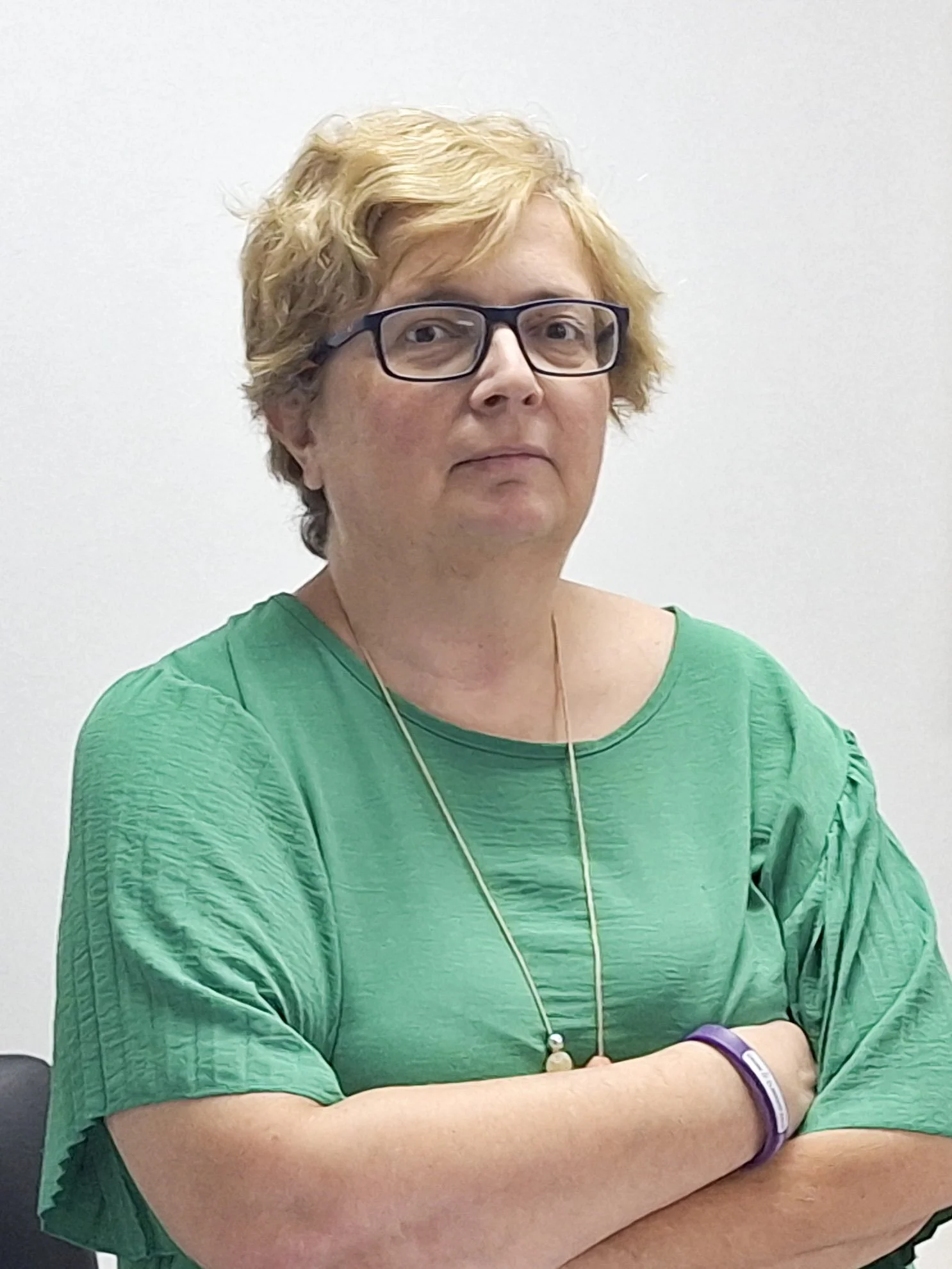 A woman with short blonde hair, black glasses, wearing a green shirt, and a purple wristband, sitting with her arms crossed in front of a plain white wall.