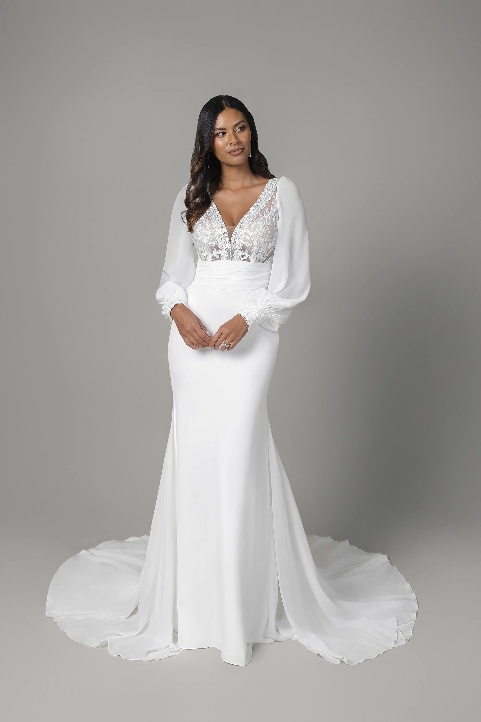 Eine Frau in einem weißen Hochzeitskleid mit langen, blousierten Ärmeln und Spitze-Details am Oberteil, vor einem grauen Hintergrund.