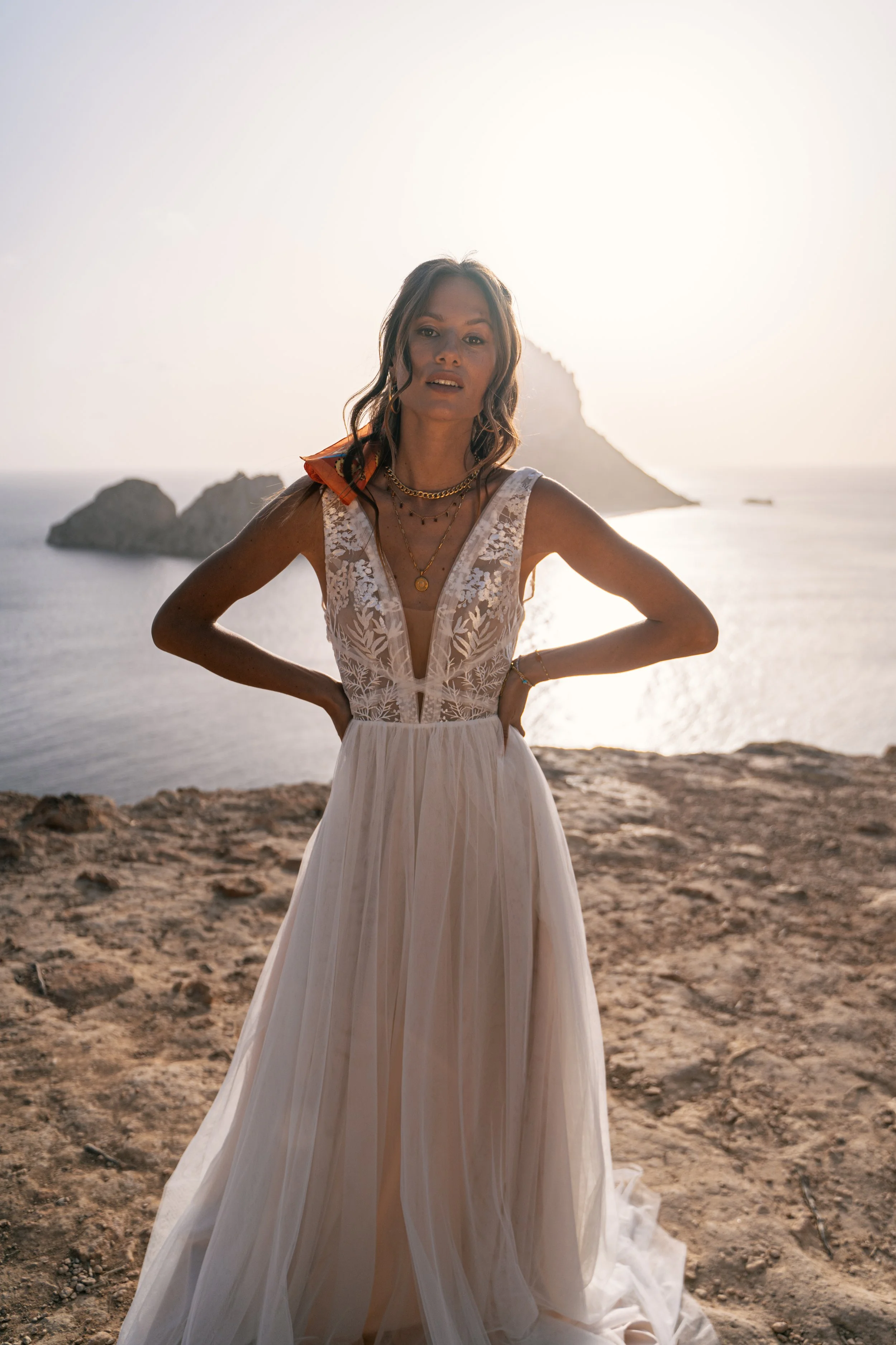 Frau in einem weißen, steifen Brautkleid mit Spitze in einer Strandkulisse bei Sonnenuntergang, im Hintergrund das Meer und eine Felseninsel.