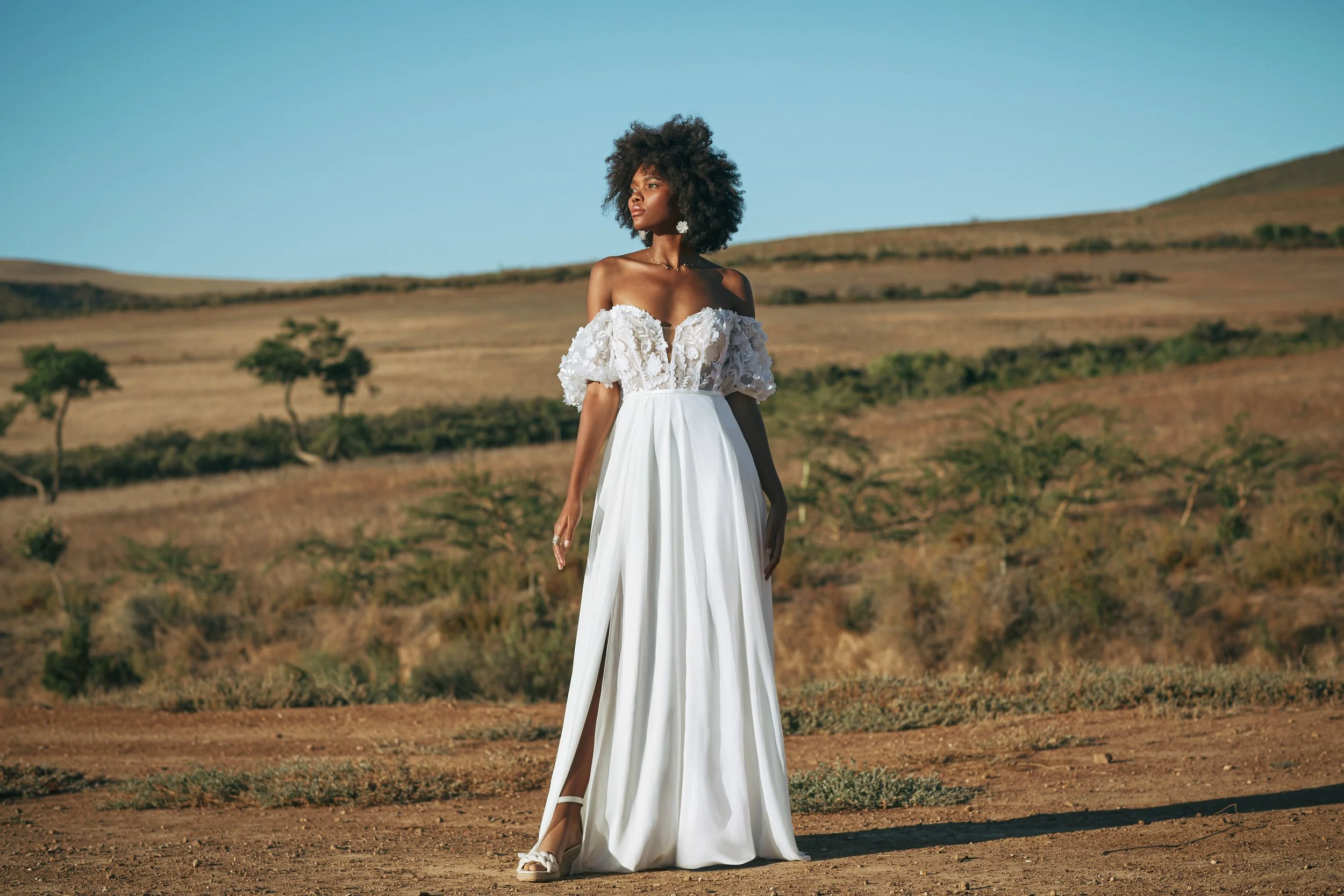 Frau in weißem Brautkleid mit Off-Shoulder-Design und Volants im offenen Land mit Hügeln und wenigen Bäumen im Hintergrund, bei Sonnenlicht fotografiert.