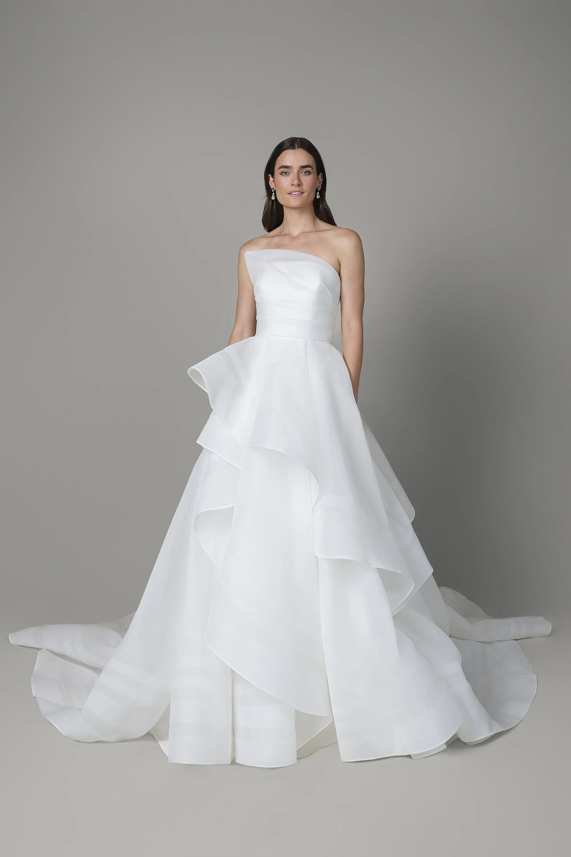 Eine junge Frau steht in einem weißen Hochzeitskleid vor einem grauen Hintergrund.