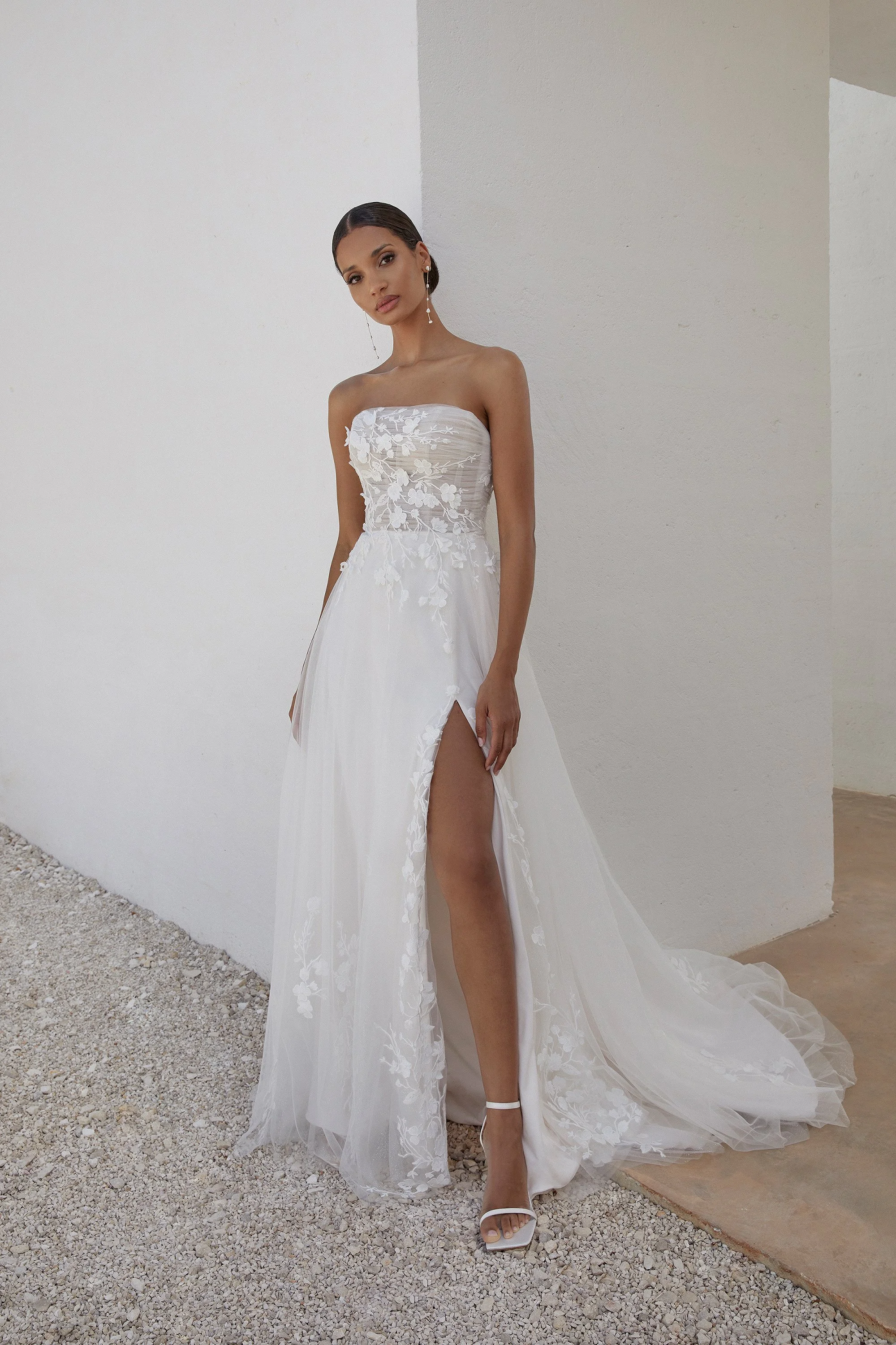 Frau in einem weißen Brautkleid mit floralem Design, das einen hohen Schlitz auf der rechten Seite hat, trägt weiße High Heels und steht auf einer Granit- und Betonfläche vor einer weißen Wand.