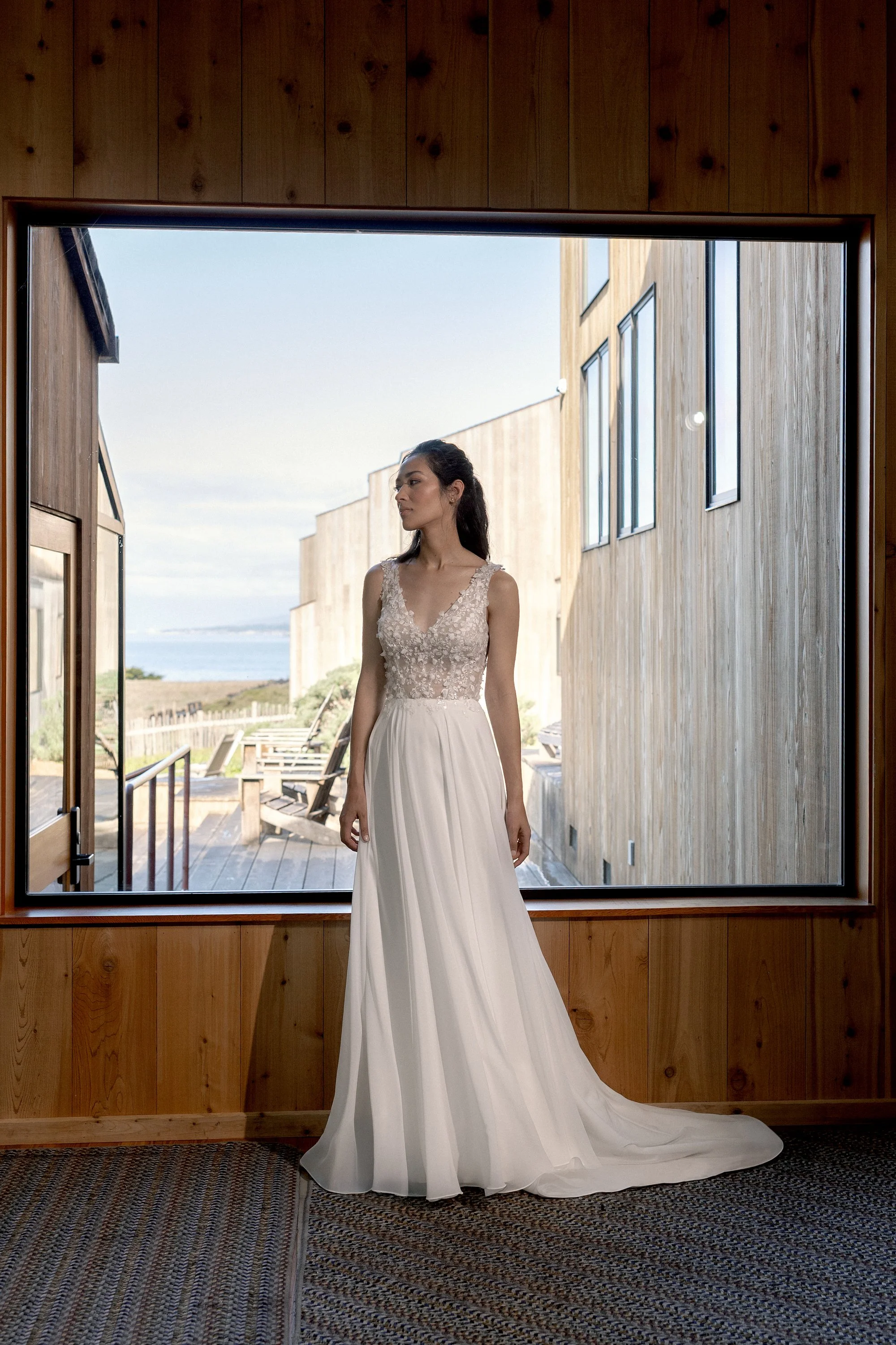 Frau im Brautkleid steht vor einem großen Fenster in einem Holzhaus, Blick nach draußen auf einen Strand und den Himmel.