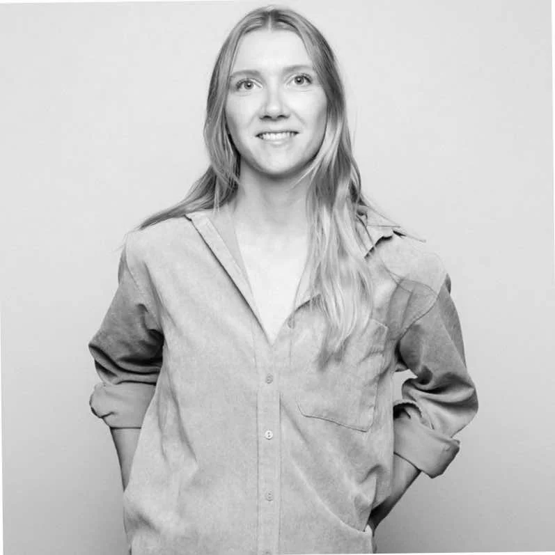 Black and white photo of a smiling woman with long hair, wearing a button-up shirt, standing with her hands behind her back.