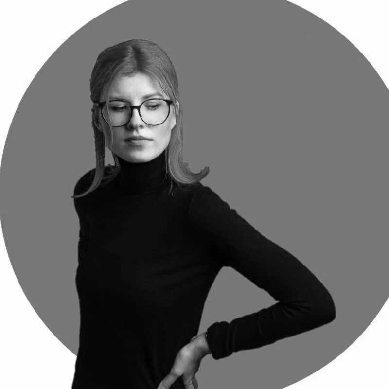 Black and white photo of a woman wearing glasses and a black turtleneck, standing with a hand on her hip.