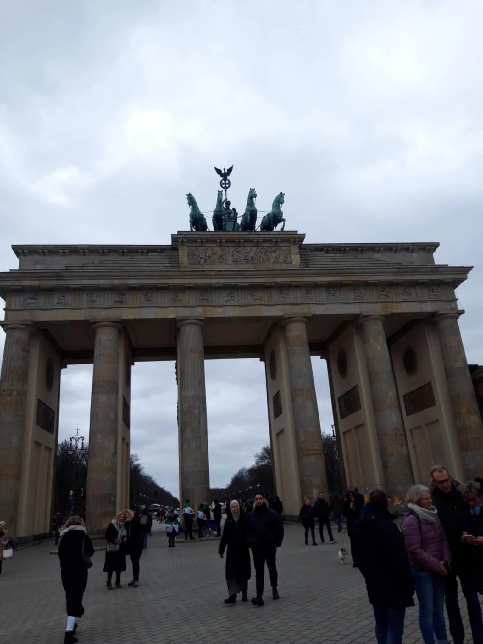 The Brandenburg Gate, a historic monument in Berlin, Germany, with people standing and walking in front of it on a cloudy day.