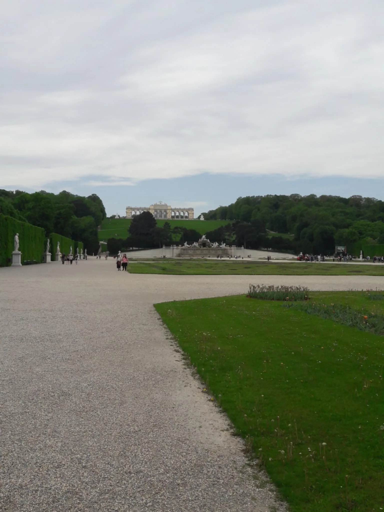 View of a formal garden with a fountain, statues, and lush green trees, leading to a grand building on a hill under a cloudy sky.