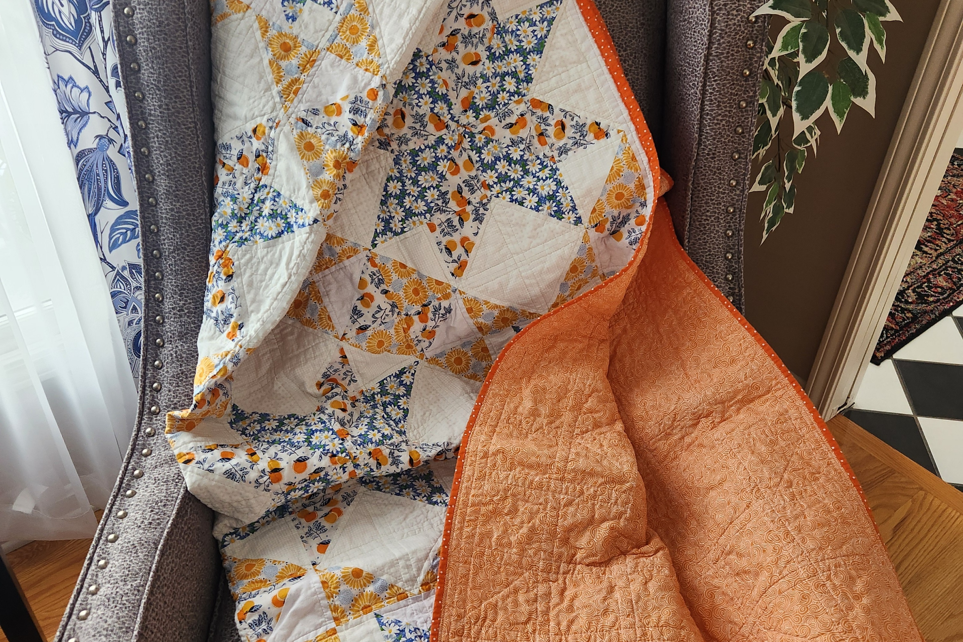 A colorful quilt with floral and geometric patterns is draped over a gray upholstered armchair. The quilt features yellow, blue, white, and orange colors with daisy and sunflower motifs. An orange fabric with a swirling pattern and orange trim is partially visible on the right side of the quilt.