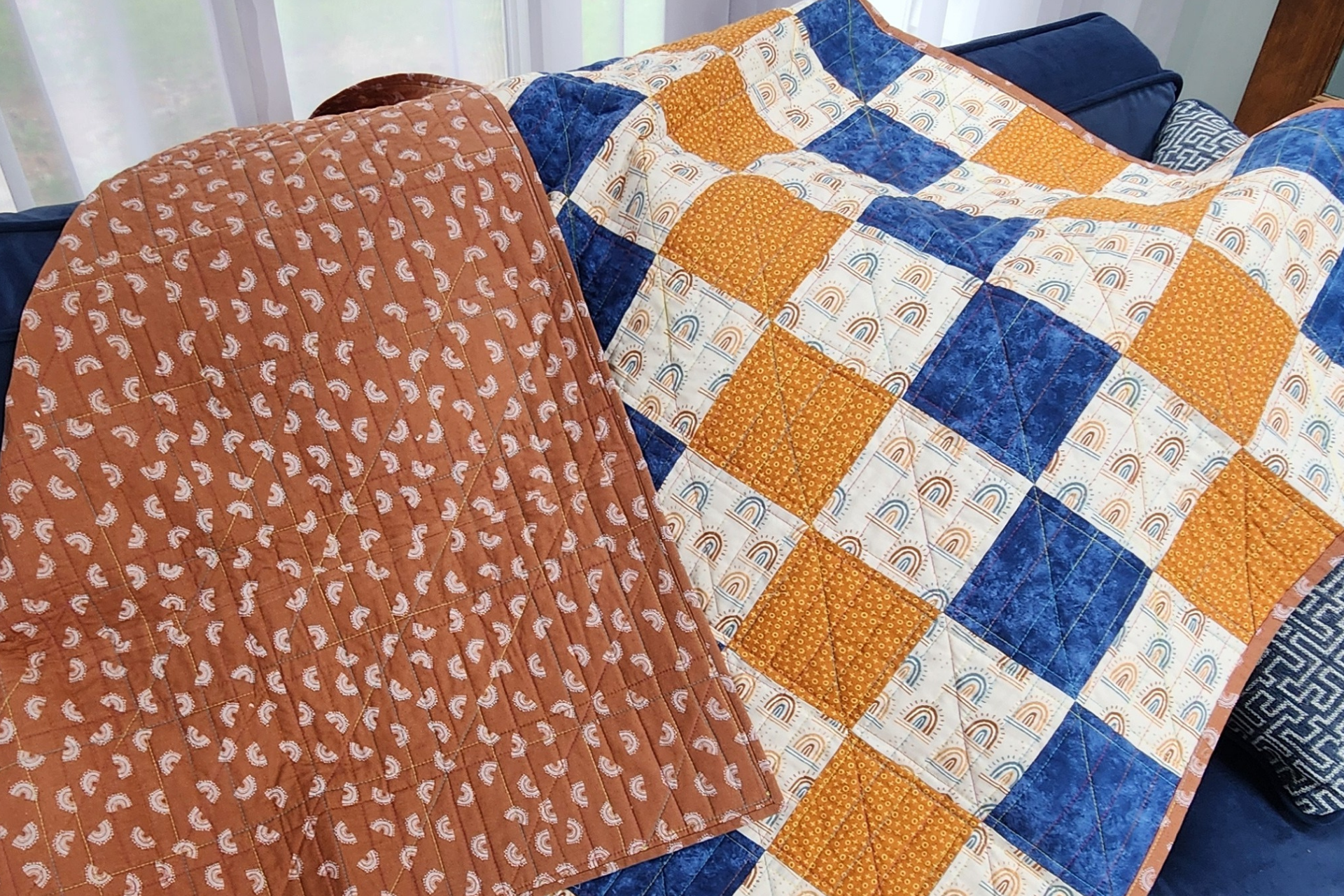 A quilt with a patchwork pattern of orange, blue, and cream colors draped over a couch, with part of the quilt turned over showing the orange backing with a white whimsical pattern.