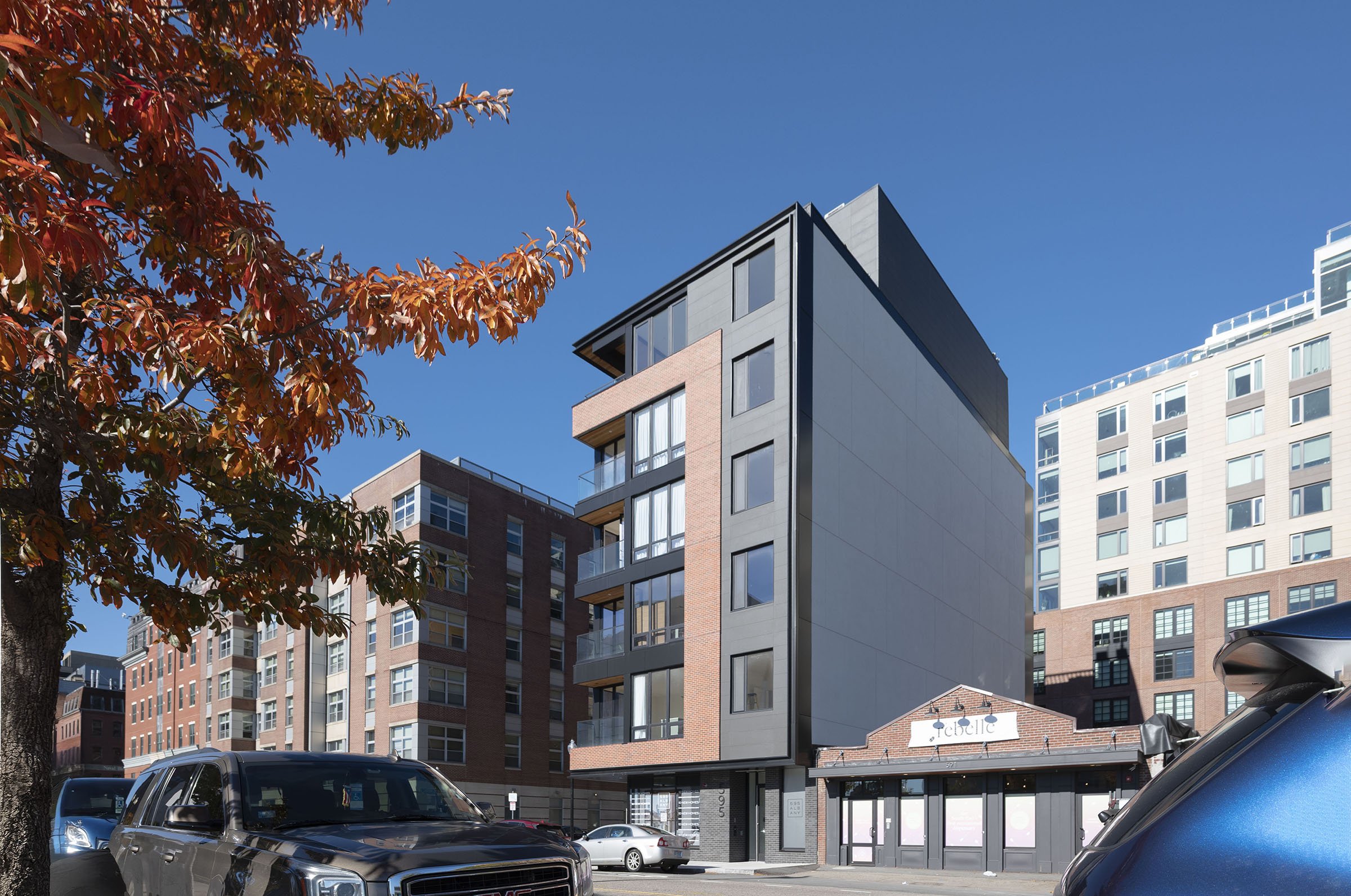Modern multi-story building with balconies and large windows, situated on a city street with parked cars and autumn-colored tree on the left, under a clear blue sky.