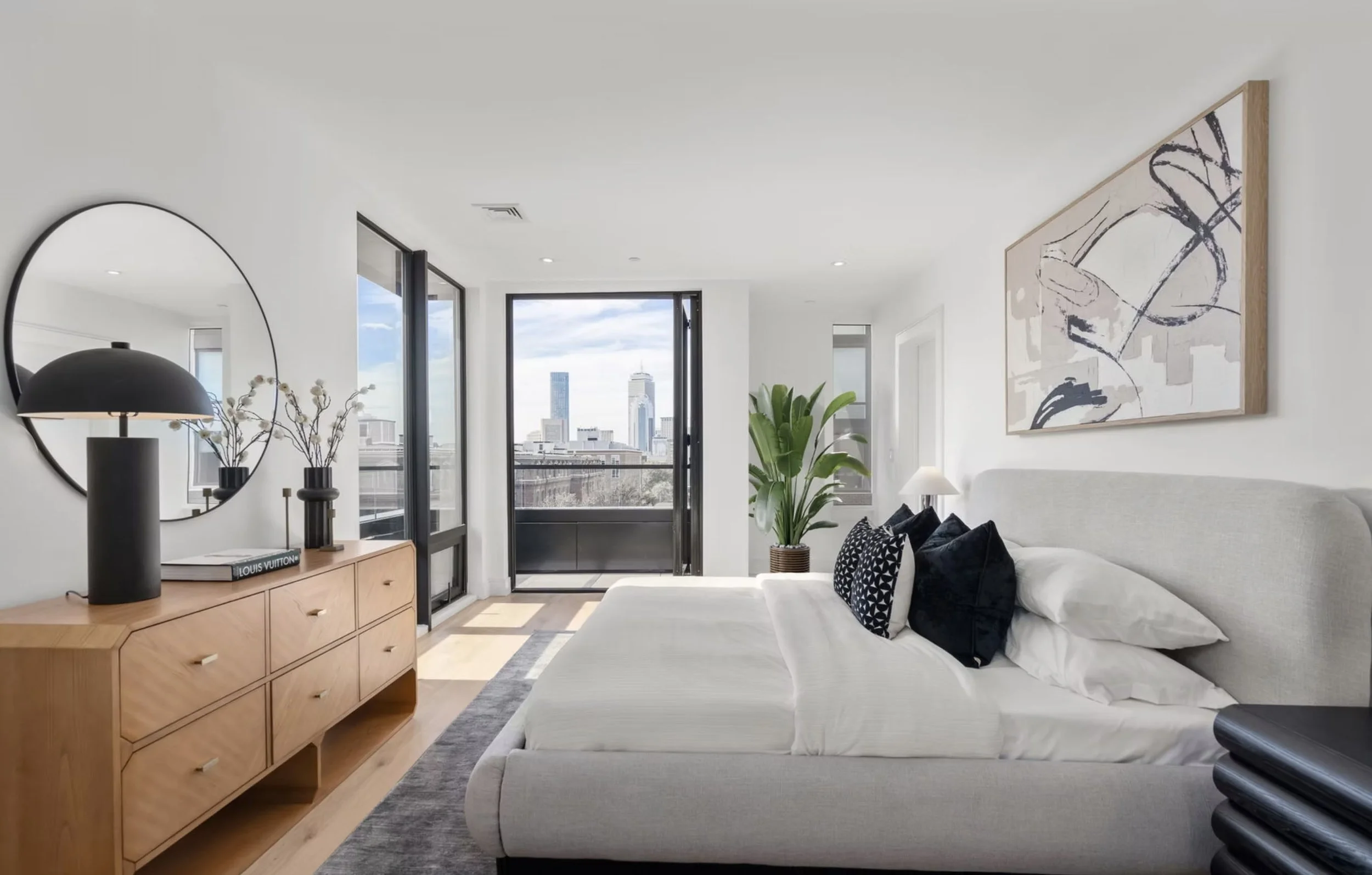 Modern bedroom with a large bed, black and white pillows, artwork above the headboard, a wooden dresser with a black lamp and books, large windows with a city view, a plant, and a balcony door.
