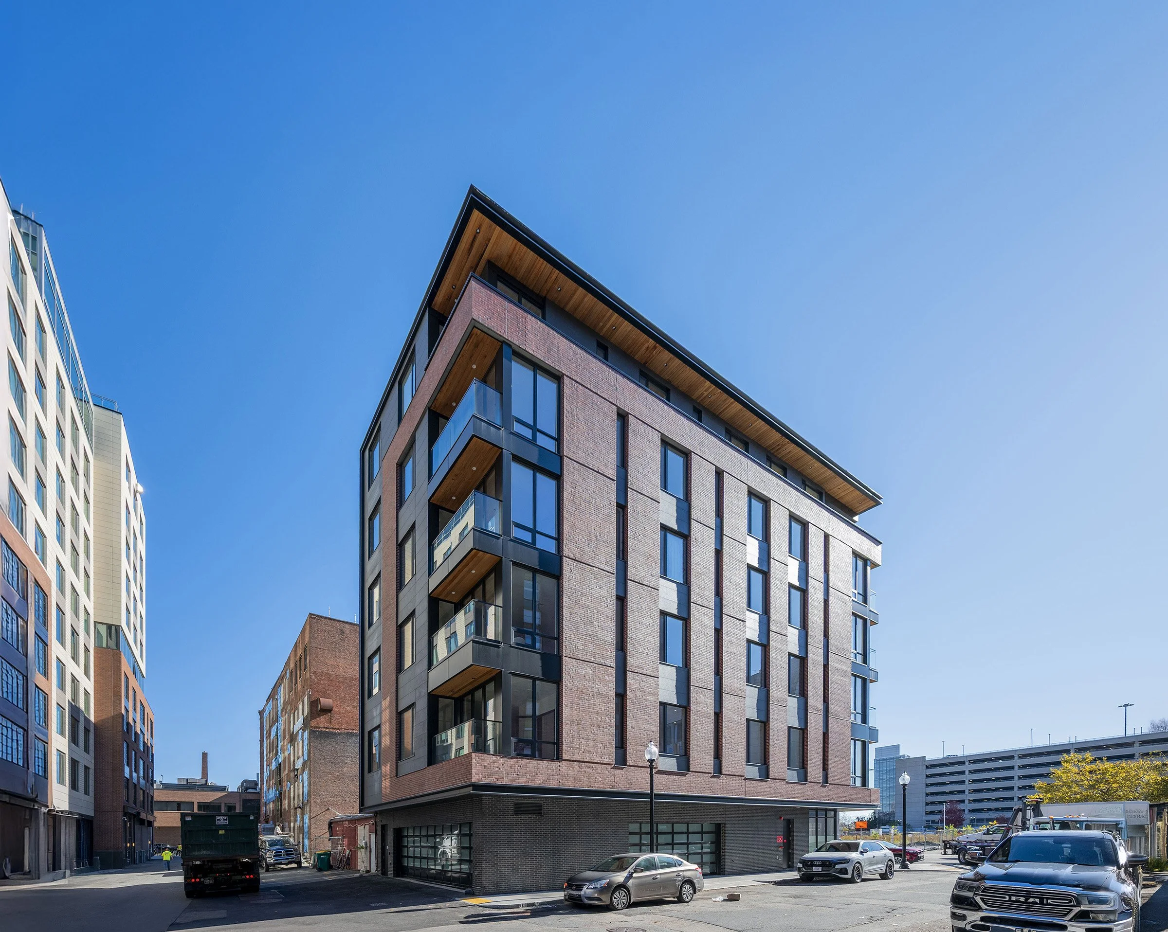 Modern multi-story brick apartment building with balconies, set against a clear blue sky, surrounded by parked cars and other urban buildings.