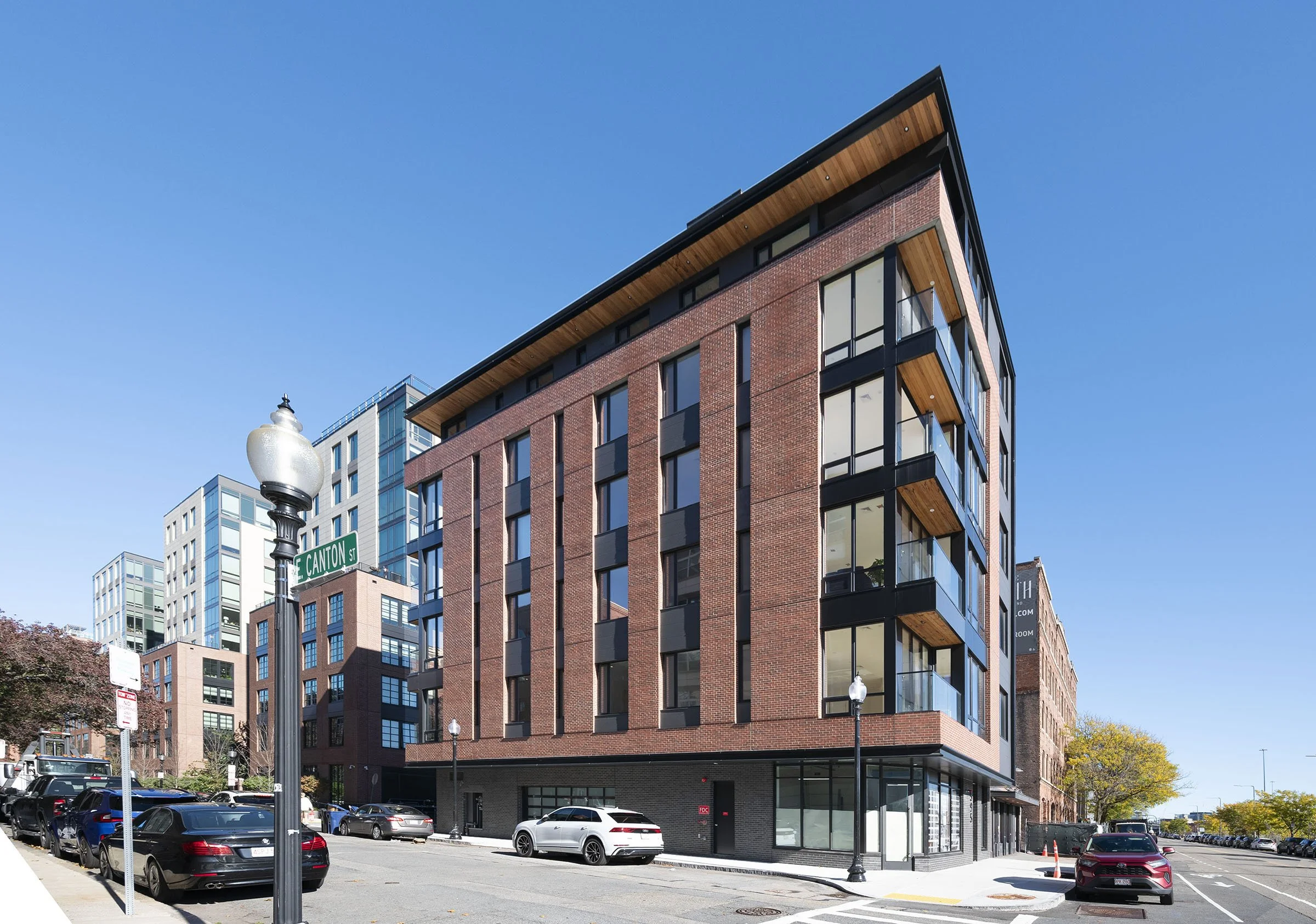 Modern multi-story red brick building with black accents and glass balconies on a city street corner under a clear blue sky, with parked cars and a street sign that reads E Canton St.