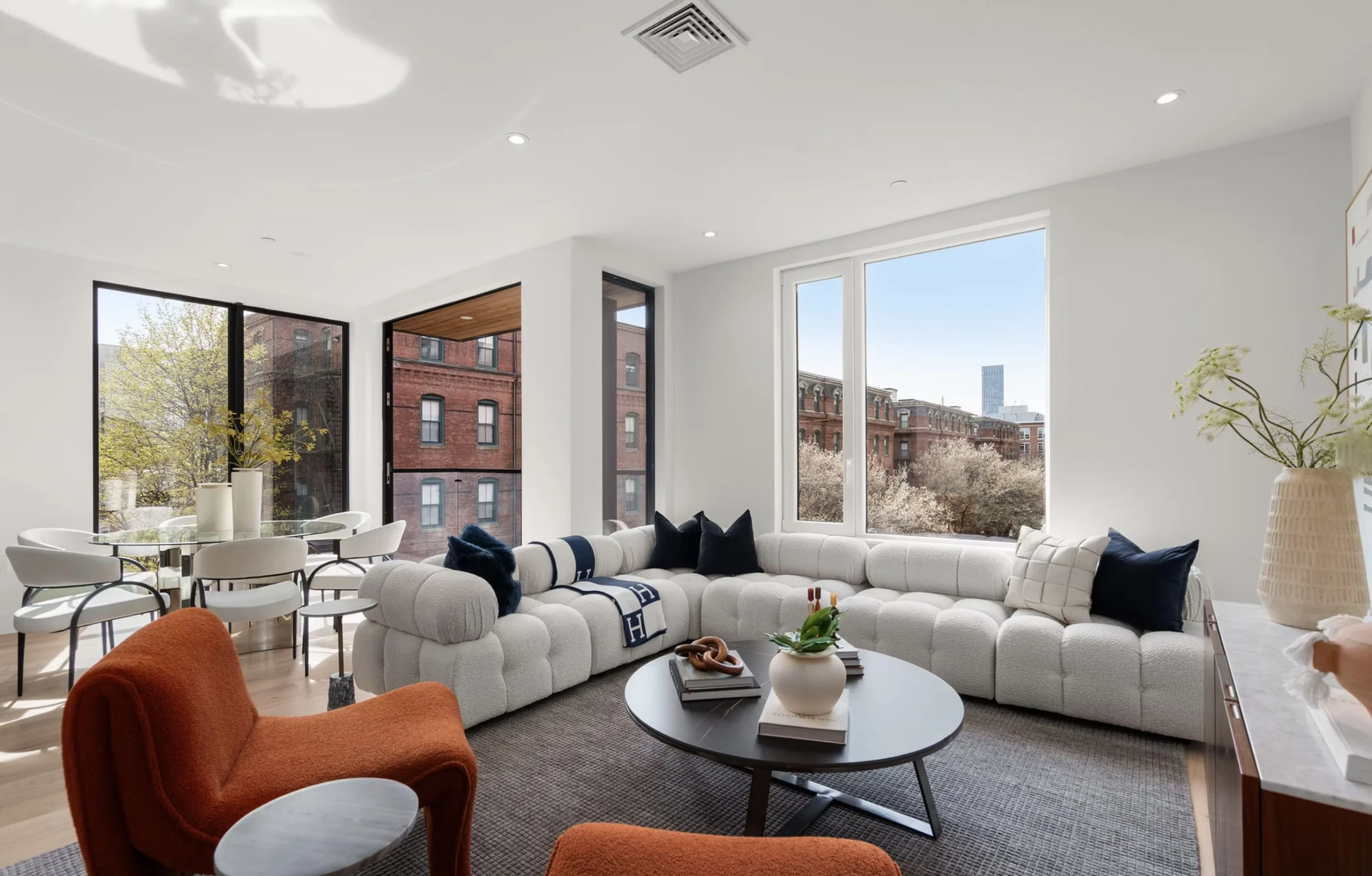 Modern living room with large windows and views of city buildings, featuring a white curved sectional sofa, navy and white accents, a round black coffee table with decor, orange armchairs, and minimalist decor.