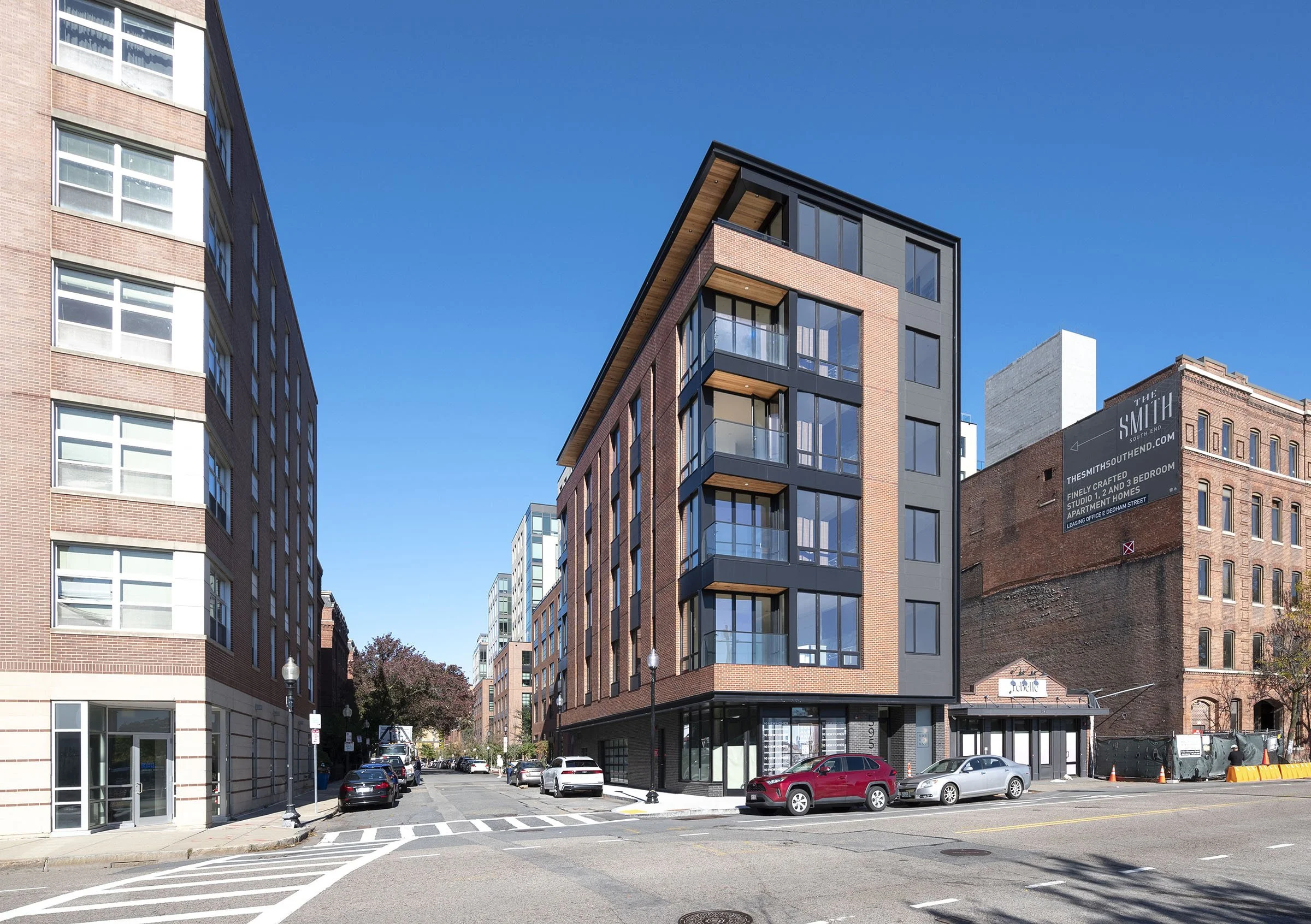 A modern multi-story brick building with large glass windows and balconies, situated on a city street with parked cars on the side.
