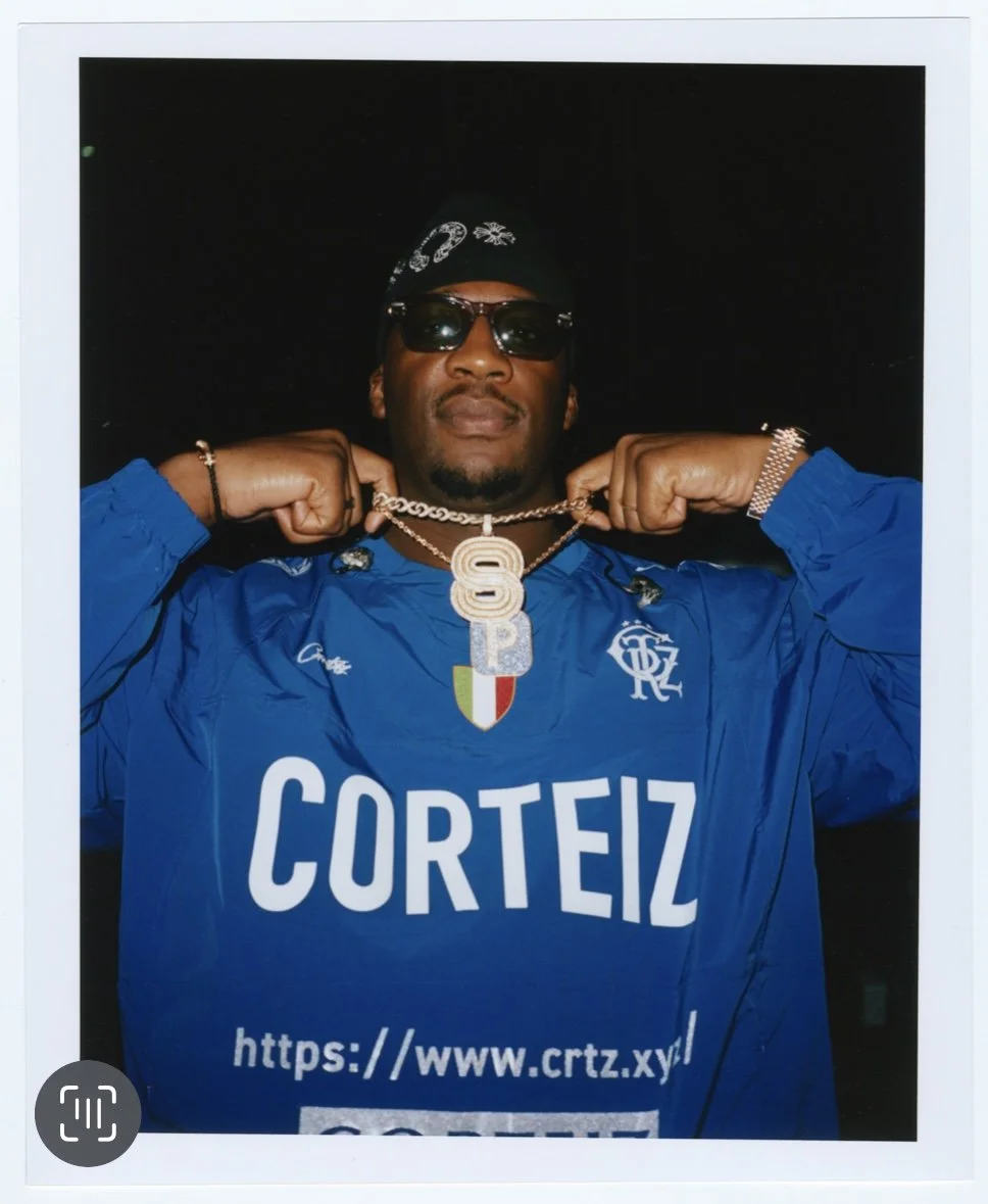 Un homme portant une veste bleue avec le mot "Corteiz" et une chaîne avec un pendentif en forme de lettre S, posant avec ses bras levés.