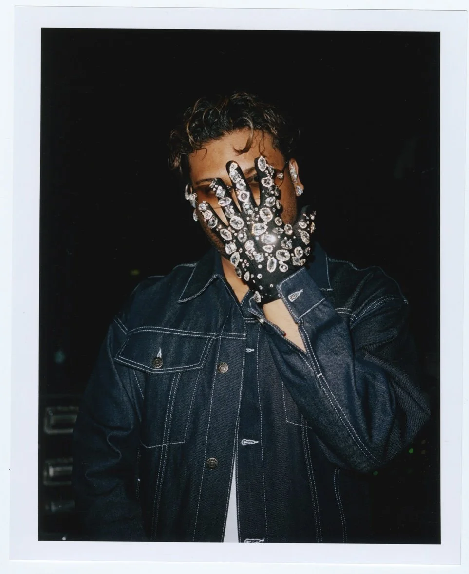 Un individu portant une veste en denim foncé, portant des gants ornés de bijoux en pierre, couvre son visage avec la main, dans un environnement sombre.