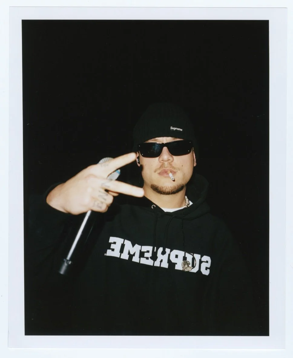 Un homme portant une casquette noire Supreme, des lunettes de soleil noires, une chemise à capuche noire avec le mot 'SOPHISTICATED' écrit dessus, tenant un cigarette et faisant un signe de la main.