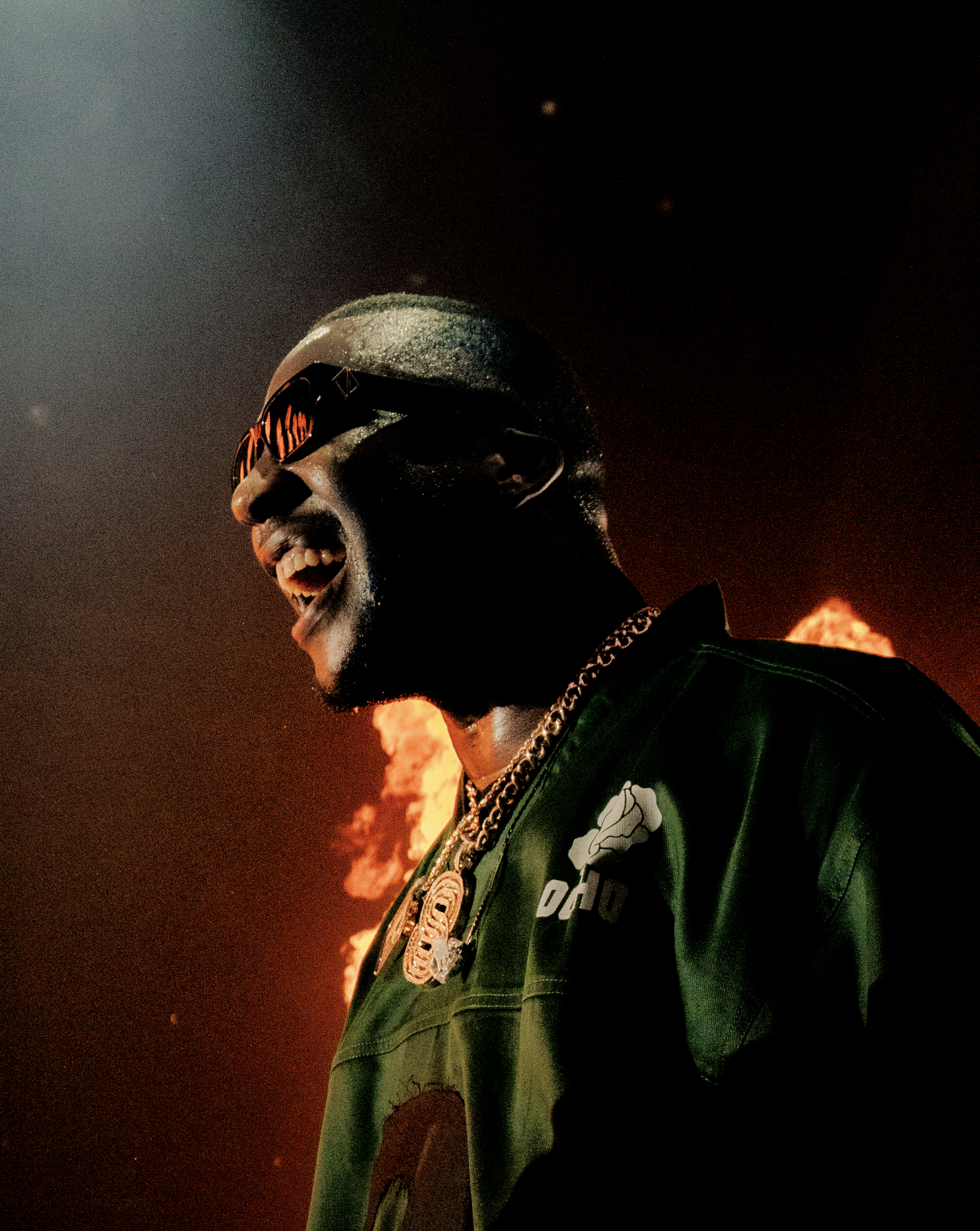 Un homme souriant portant des lunettes de soleil et une chaîne en or, avec un feu en arrière-plan dans un ciel sombre.