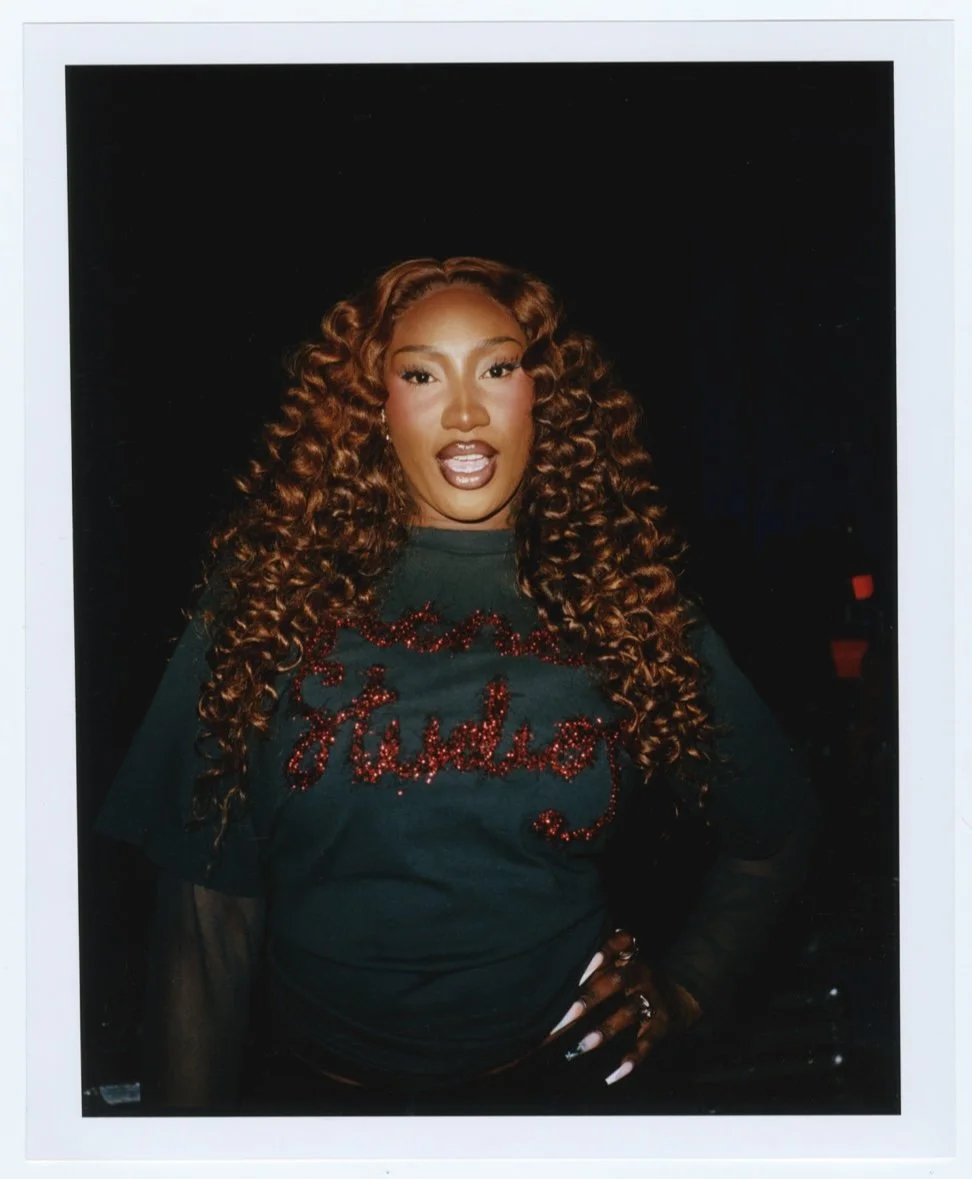 Une femme avec de longues boucles brunes, portant un t-shirt noir avec la phrase 'Glamour' en lettres rouges pailletées, pose contre un fond noir.