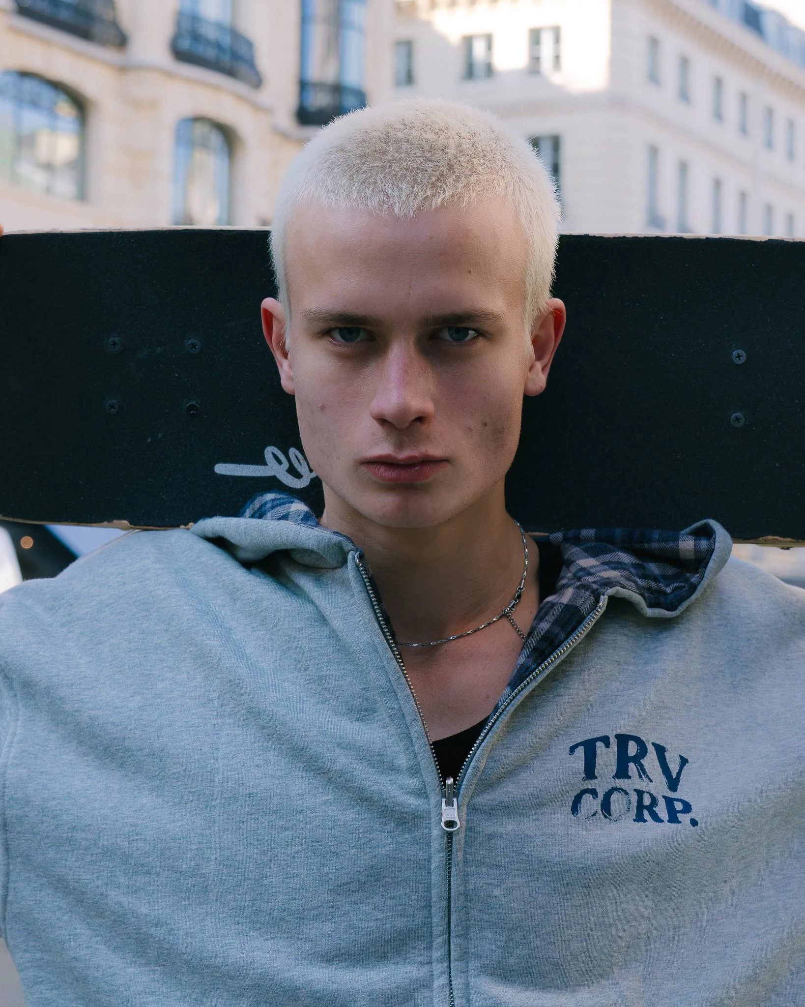 Jeune homme aux cheveux blonds courts, portant une veste grise avec logo 'TRV CORP', portant un collier, avec un skateboard sur ses épaules, en milieu urbain.