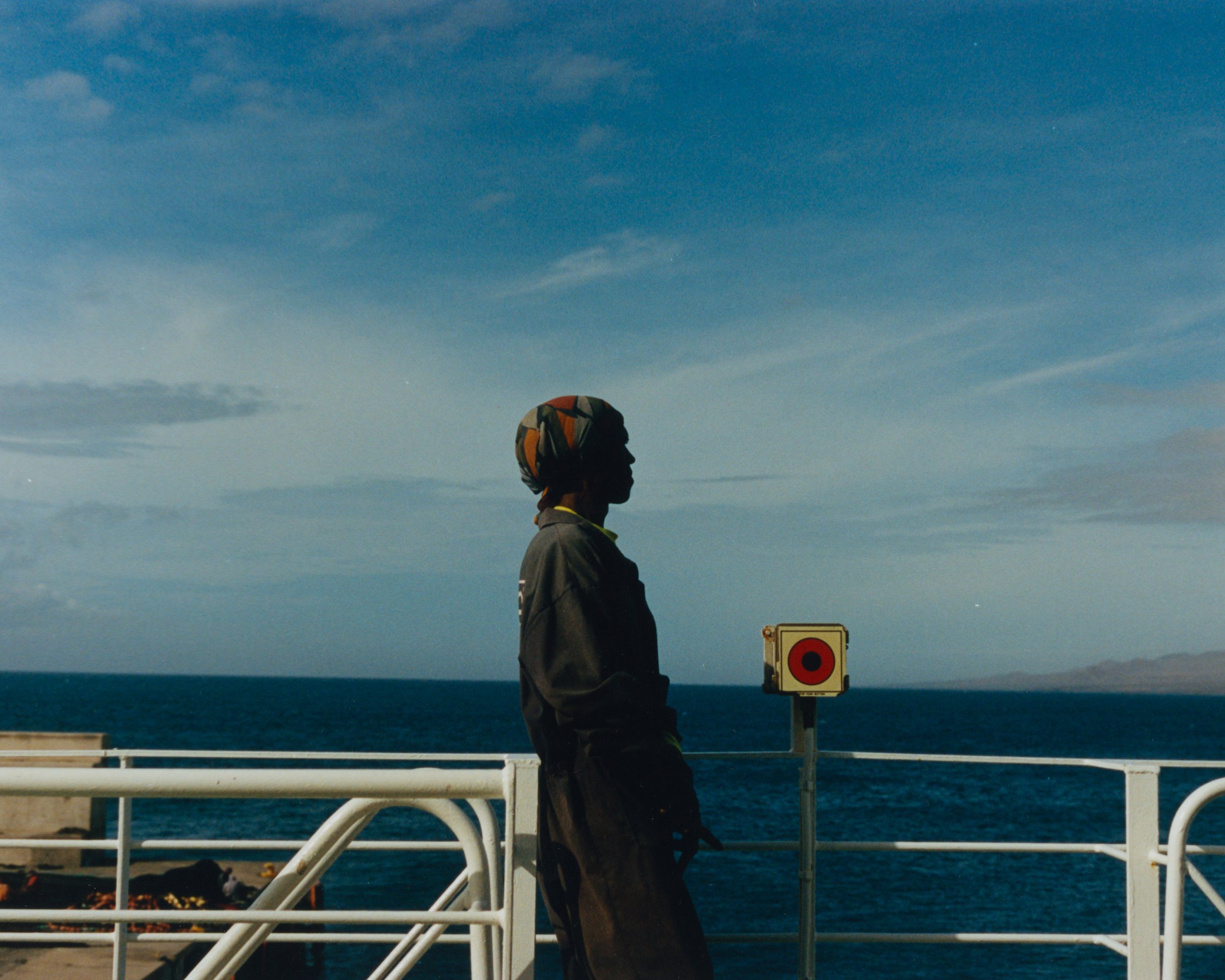 Un homme en tenue sombre, portant un casque coloré, se tient sur le pont d'un bateau face à l'océan, avec un ciel bleu clair et des nuages légers.