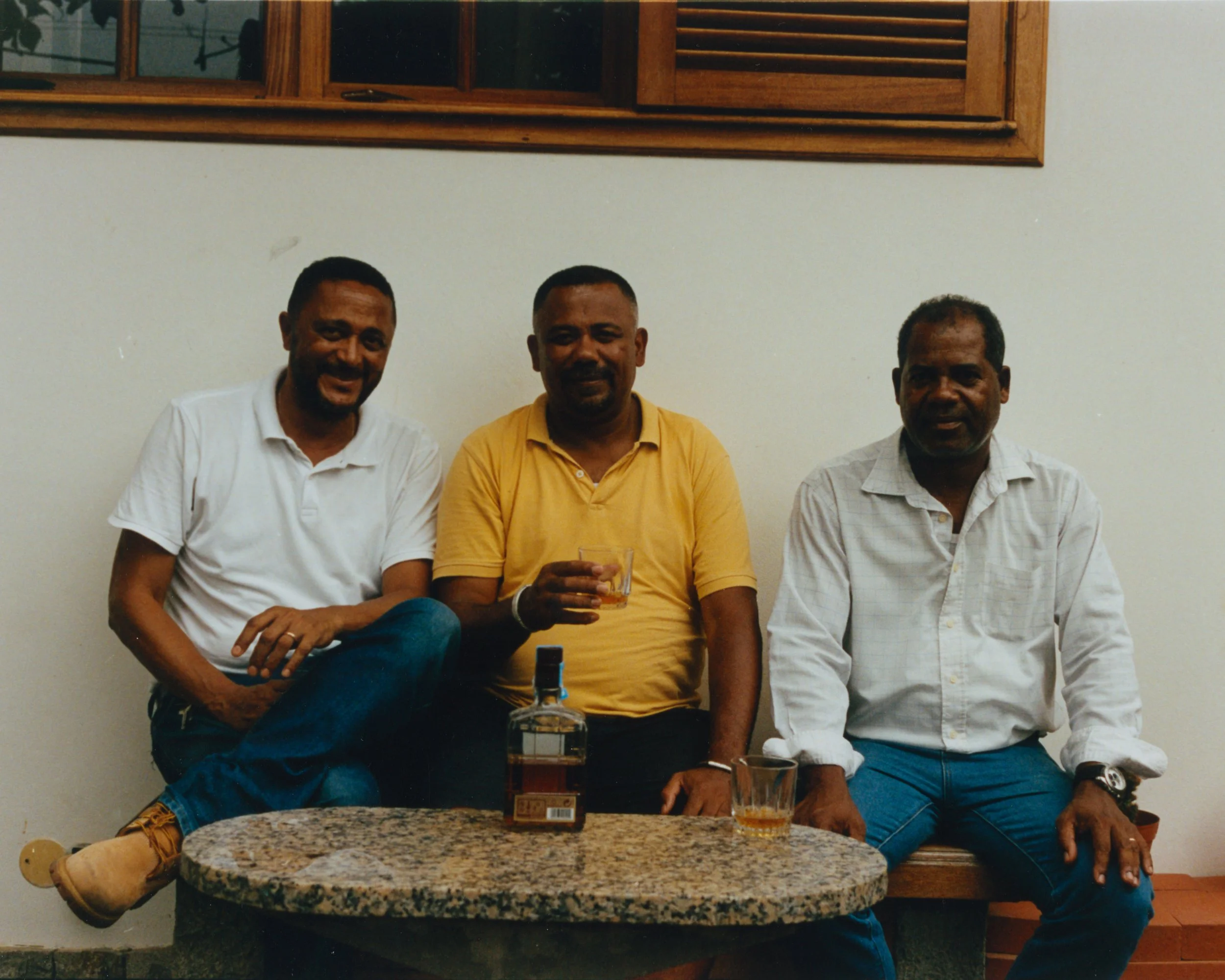 Trois hommes assis sur un banc en bois derrière une table en pierre, avec une bouteille et deux verres, posés sur la table. Un mur blanc avec une fenêtre en bois derrière eux.