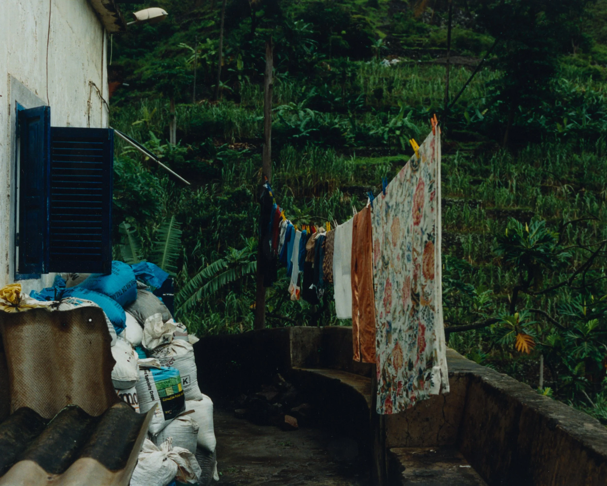 Linge suspendu pour sécher des vêtements, dans un environnement rural avec une maison en pierre, des sacs de stockage et une végétation dense en arrière-plan.
