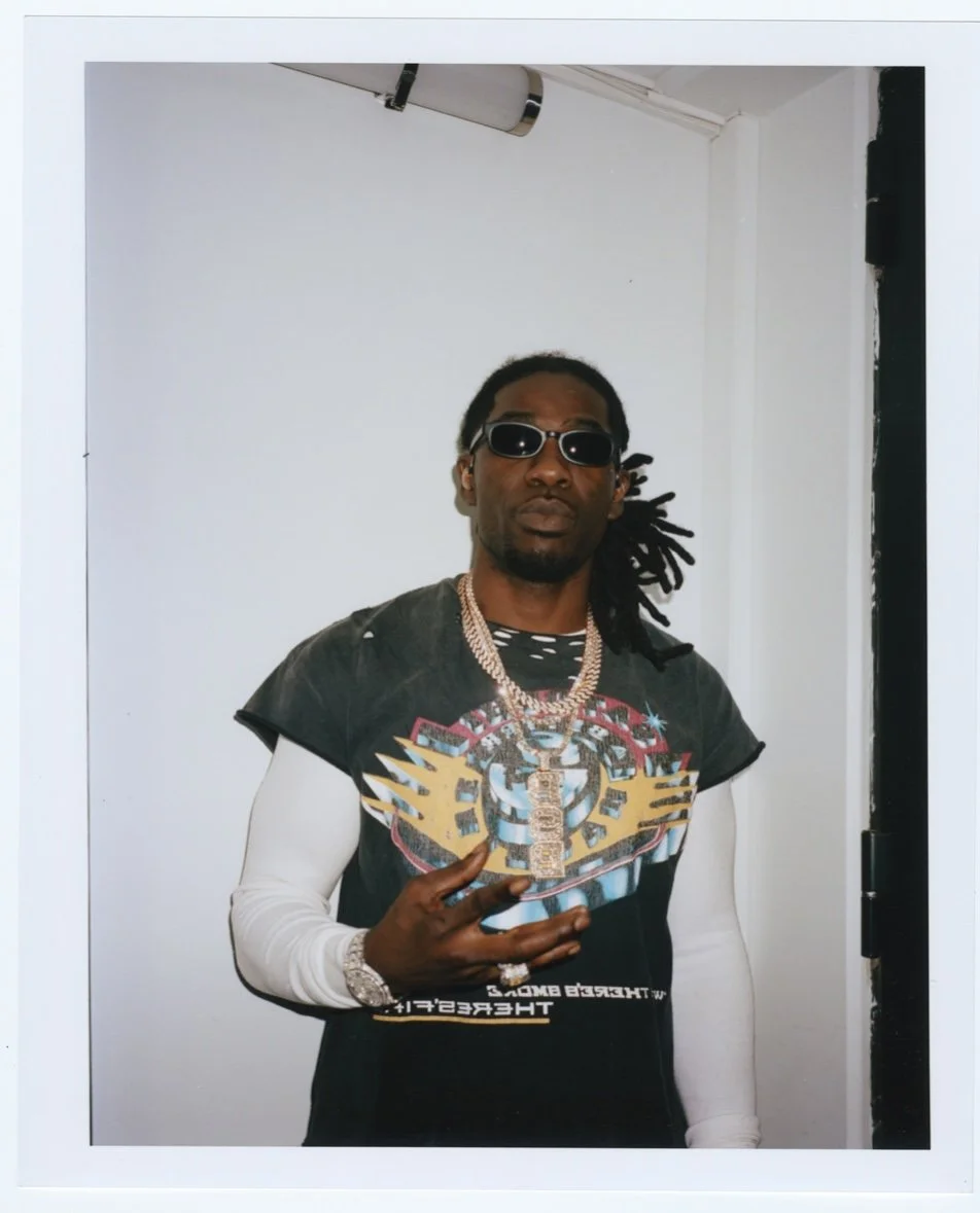 Un homme portant des lunettes de soleil, un t-shirt graphique, plusieurs colliers en or et une montre, posant devant un mur blanc.