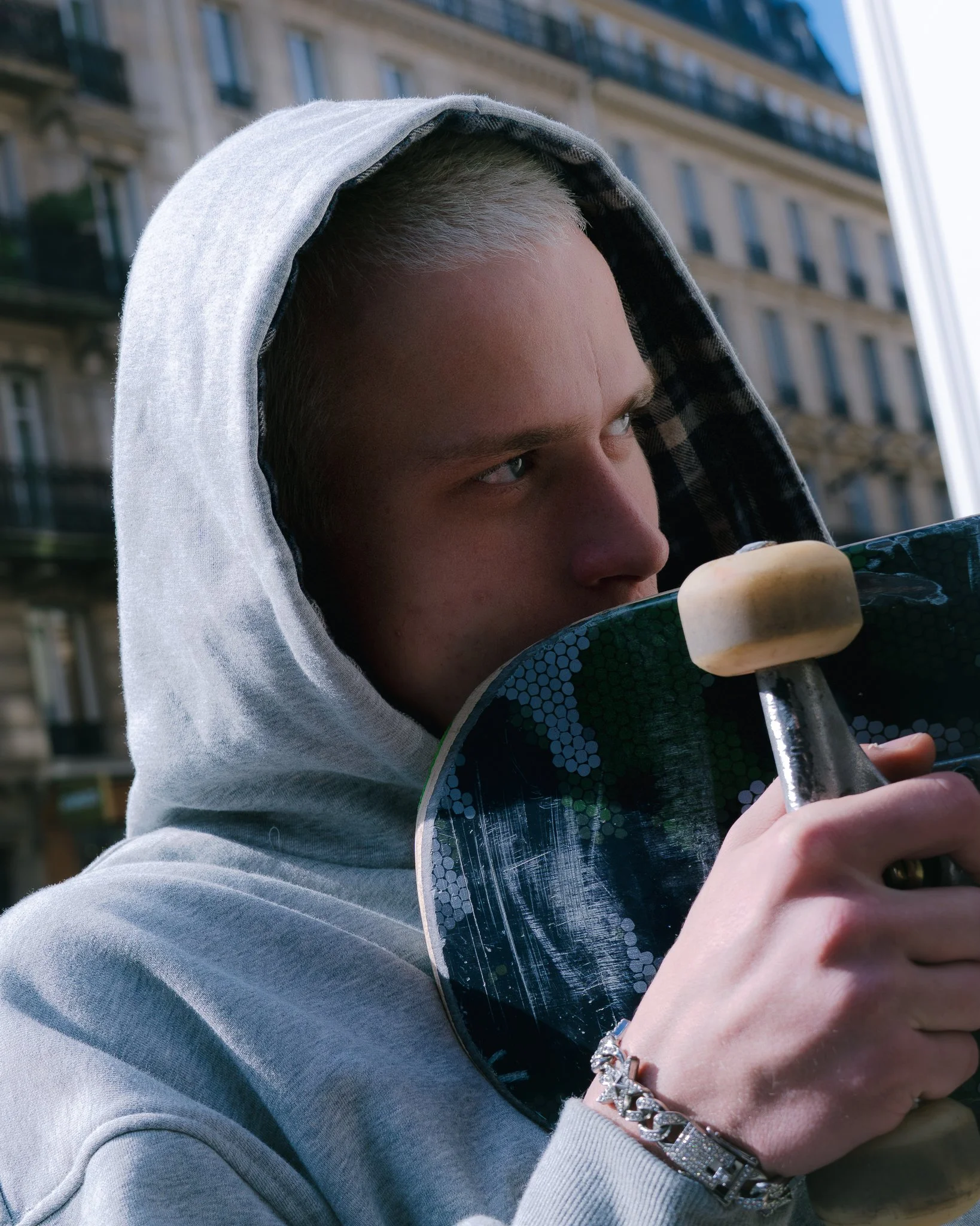 Jeune homme avec une veste à capuche gris clair tenant une planche de skateboard avec une roue blanche, en milieu urbain.