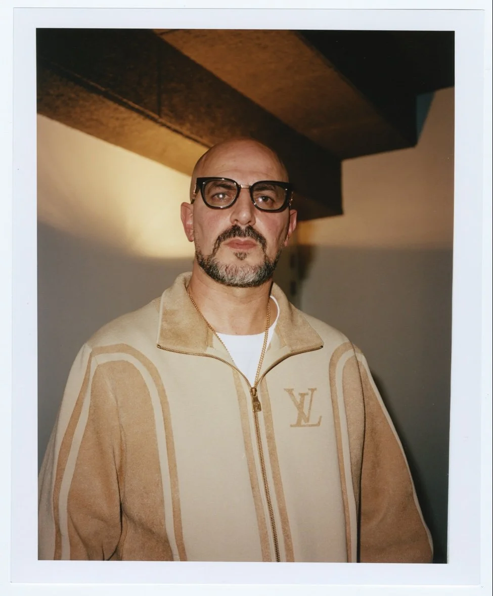 Homme portant des lunettes noires et une veste beige avec un logo LV, posé devant un mur intérieur.