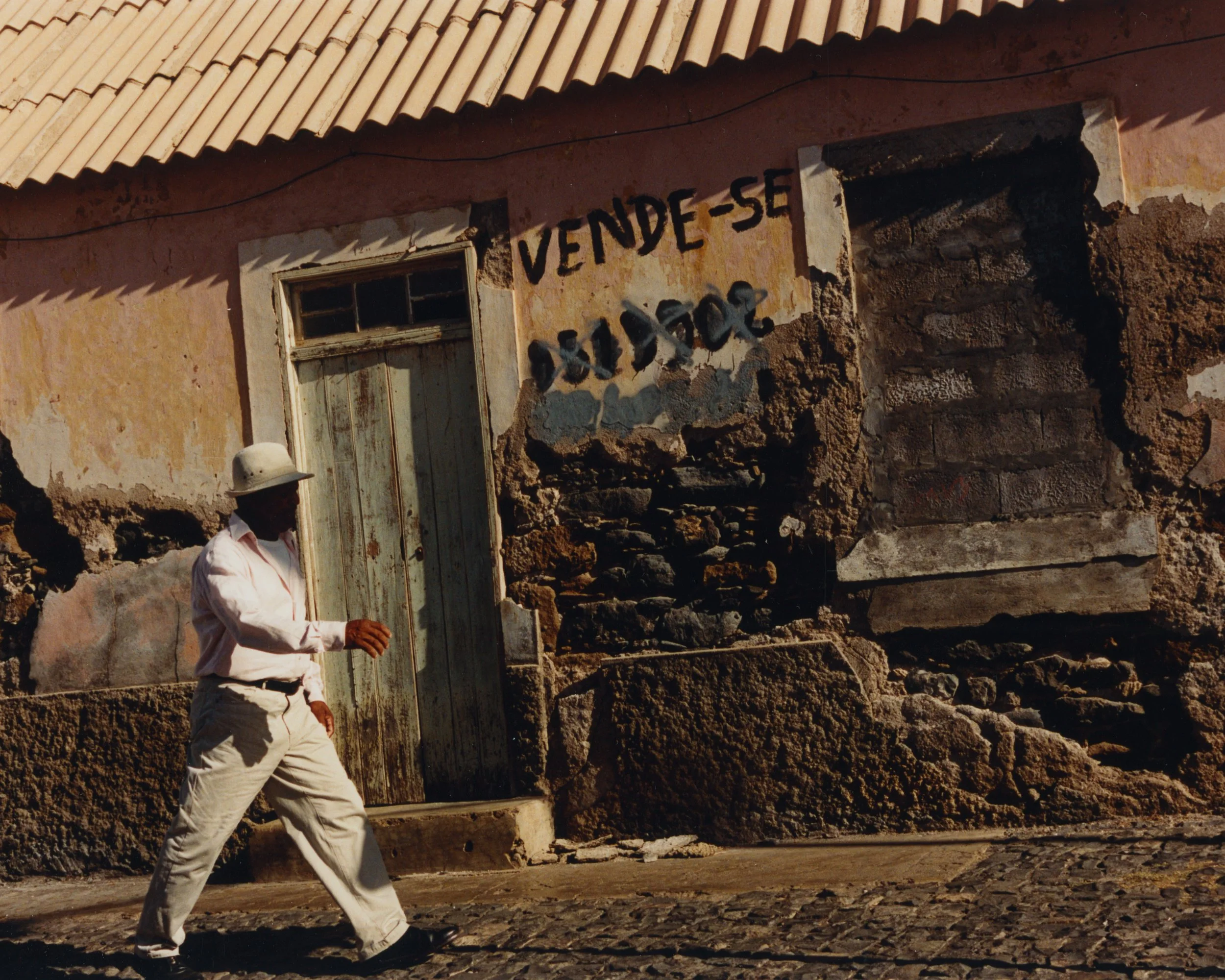 Une personne marchant devant une maison ancienne en ruines avec une affiche où il est écrit 'VEND-SE' et d'autres mots effacés, en plein jour.