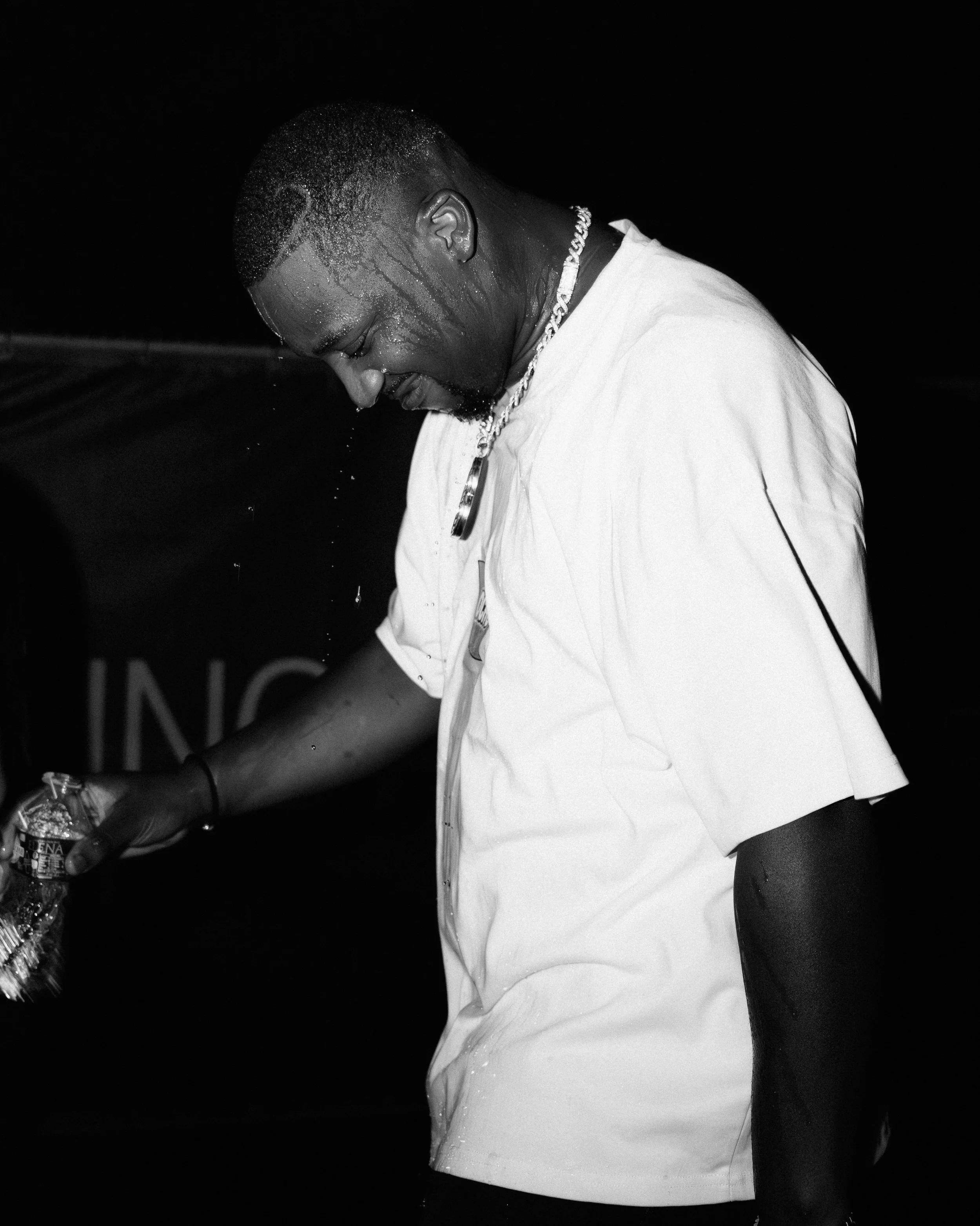 Un homme souriant, portant un t-shirt blanc, qui se rince la tête avec de l'eau dans un environnement sombre.