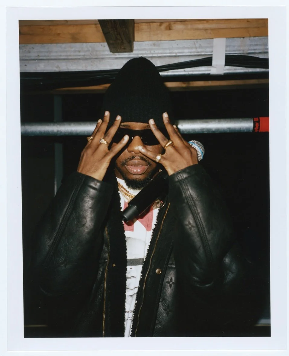 Personne portant une cagoule noire, lunettes de soleil, veste en cuir, et plusieurs bijoux. Elle couvre partiellement son visage avec ses mains, dans un environnement intérieur avec un plafond en bois et des câbles électriques.