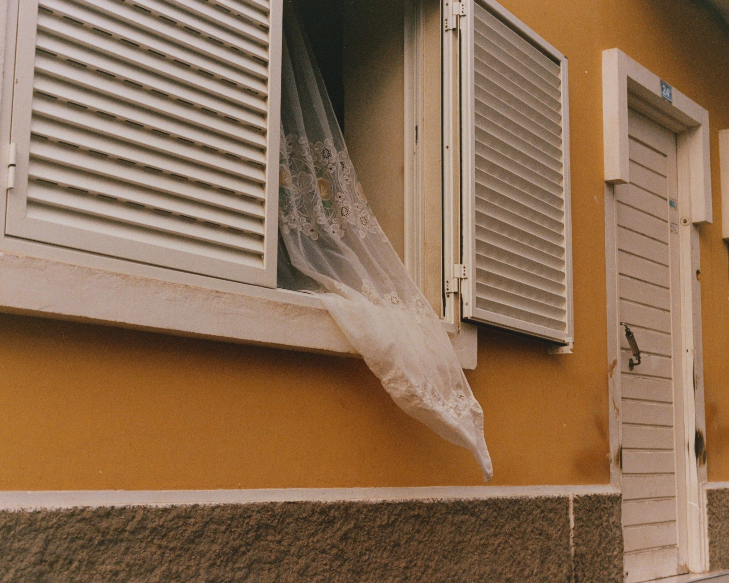 Fenêtre ouverte avec une façade en jaune et volets blancs, avec un rideau blanc qui dépasse du volet gauche.