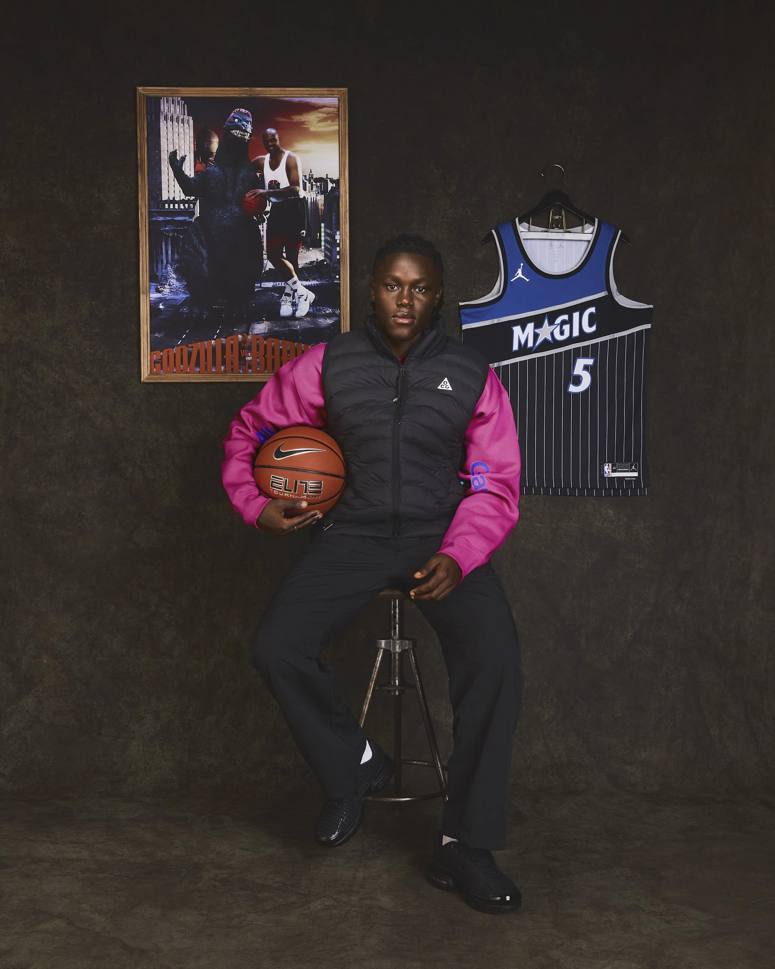 Un jeune homme noir assis sur un tabouret, tenant un ballon de basketball Nike, portant une veste noire avec manches roses et un pantalon noir, dans une pièce avec un poster de Godzilla vs. Kong et un maillot de basketball Orlando Magic au mur.