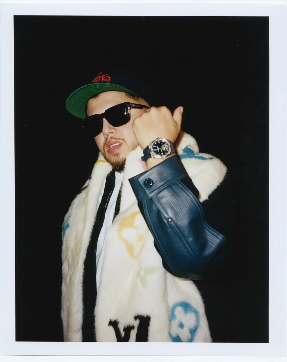 Un homme portant une casquette verte, des lunettes de soleil noires, une montre sophistiquée, et un manteau blanc avec des motifs colorés, pose avec une ambiance de mode.