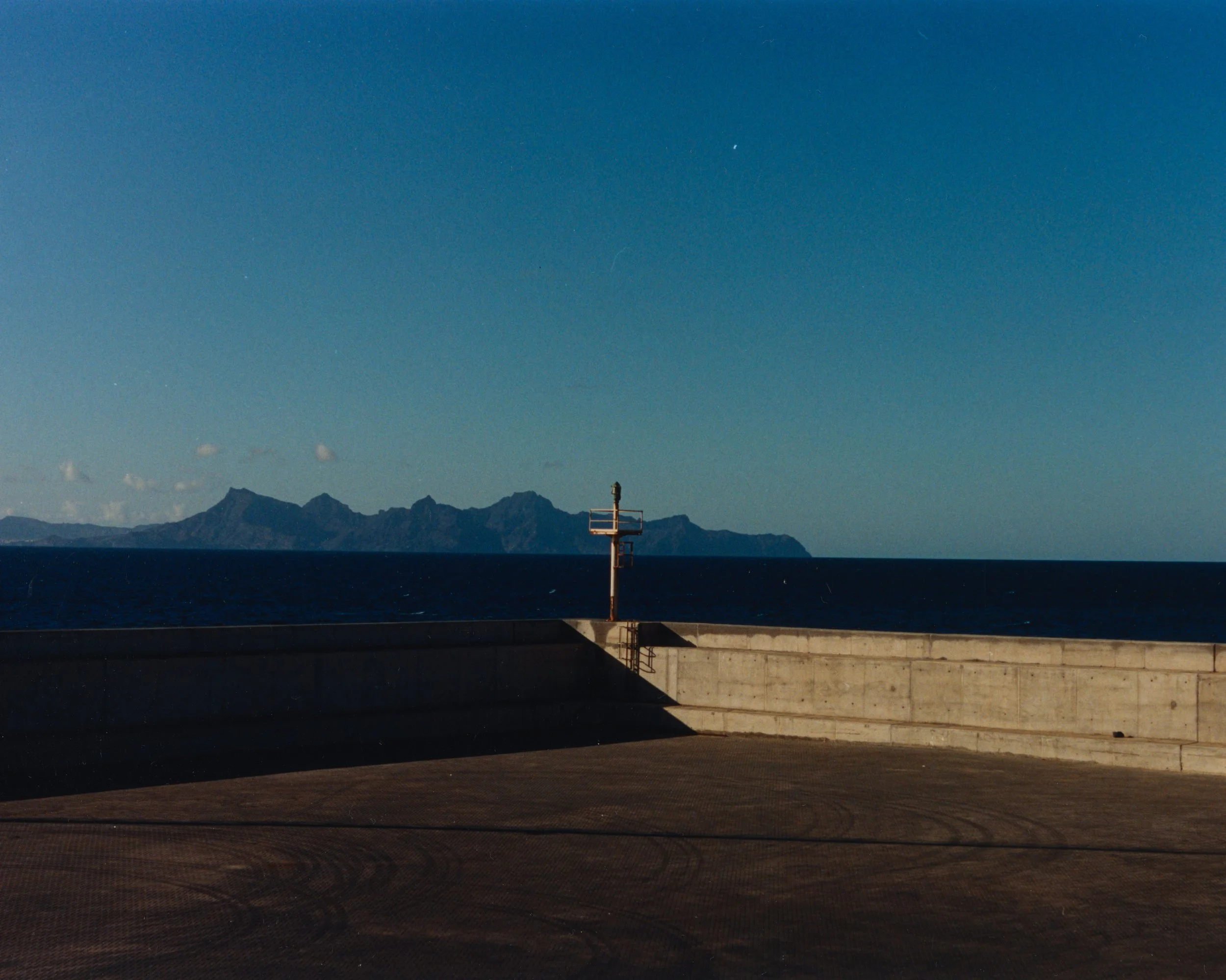 Un quai en béton face à la mer, avec un mur de protection, un phare en métal et une montgolfière en arrière-plan avec des montagnes à l'horizon.