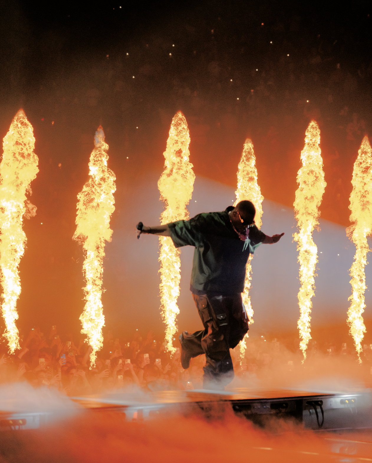 Un artiste de scène danse devant des jets de feu pendant un spectacle nocturne avec un public visible en arrière-plan.