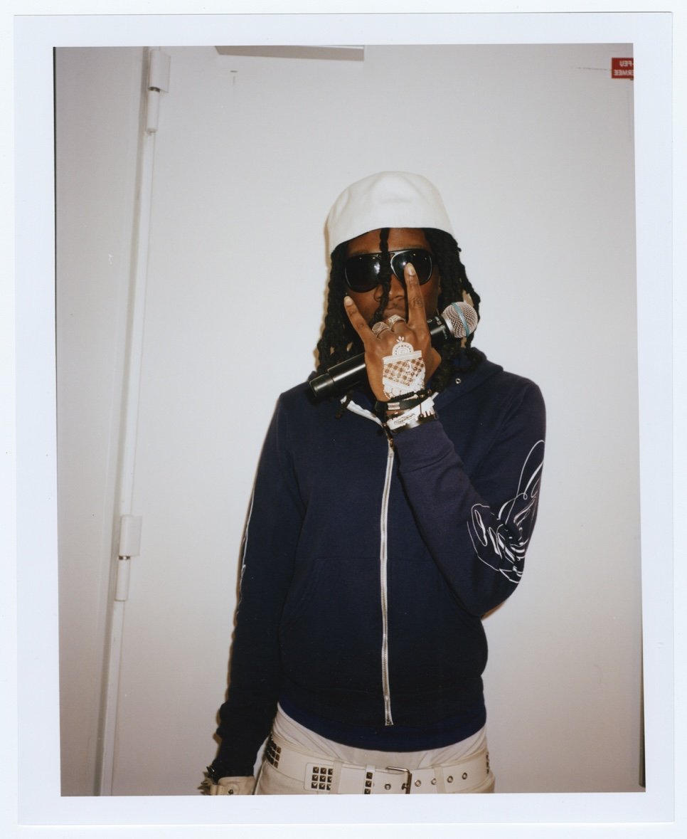 Personne portant des lunettes de soleil, un chapeau blanc, une veste sombre avec une citation en français sur la manche, tenant un microphone, faisant un geste avec la main en face de son visage, contre un mur blanc.