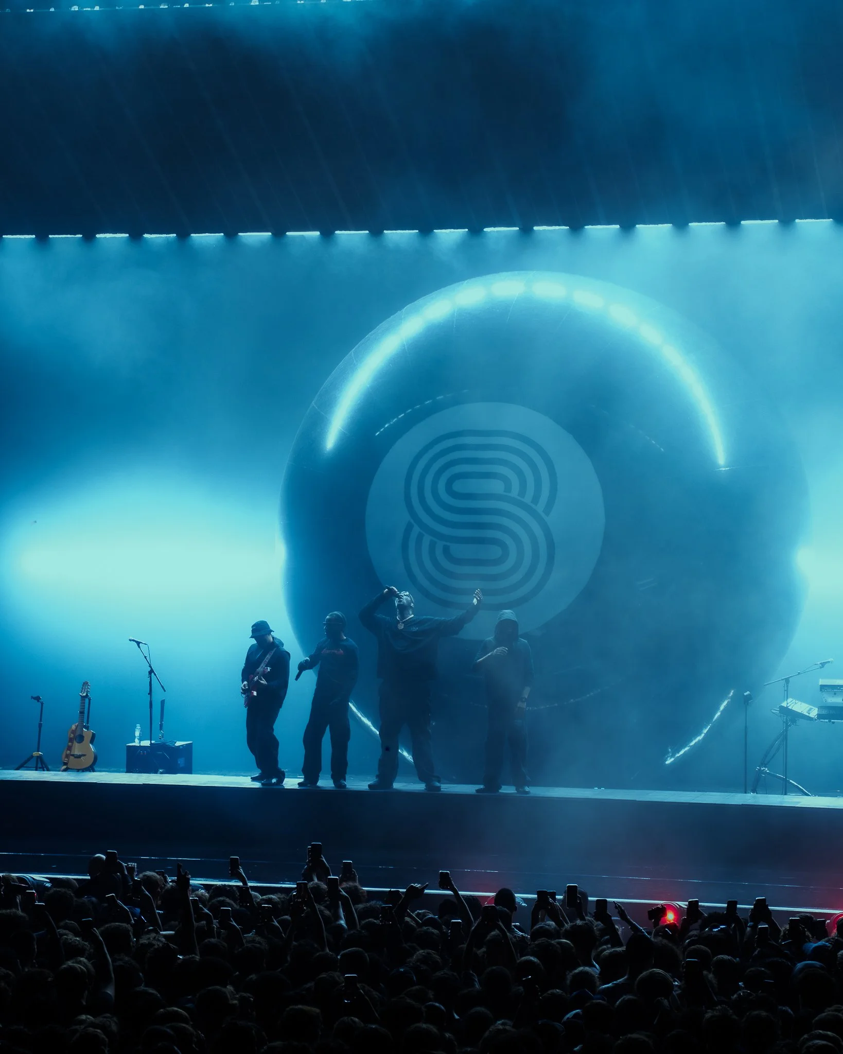 Un groupe de musiciens sur scène lors d'un concert, avec un grand écran en arrière-plan affichant un logo en forme de S à l'intérieur d'une boule, éclairage bleu et foule au premier plan