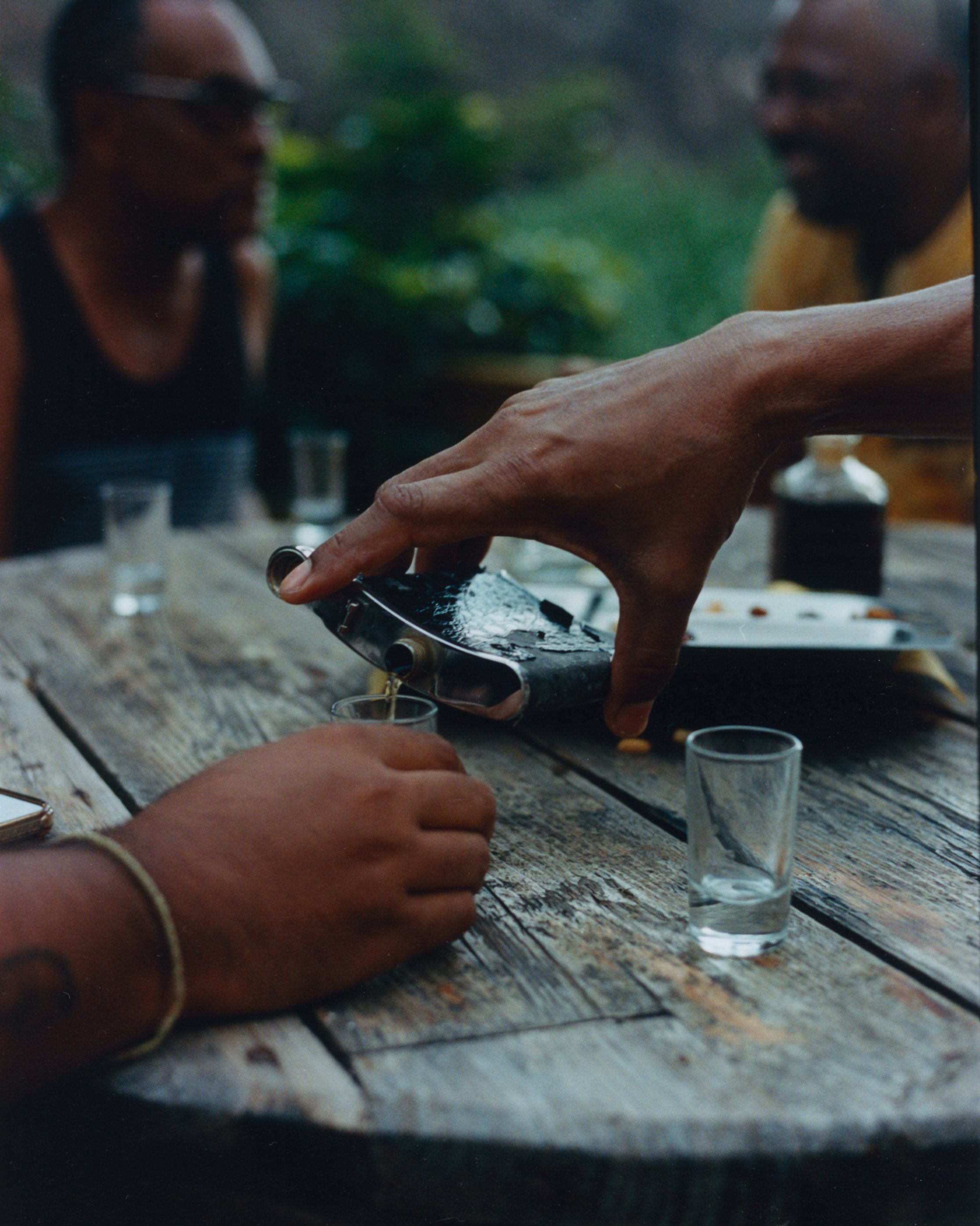 Groupe de personnes partageant un moment convivial autour d'une table en bois, avec un verre d'alcool et des snacks, dans un environnement extérieur verdoyant.