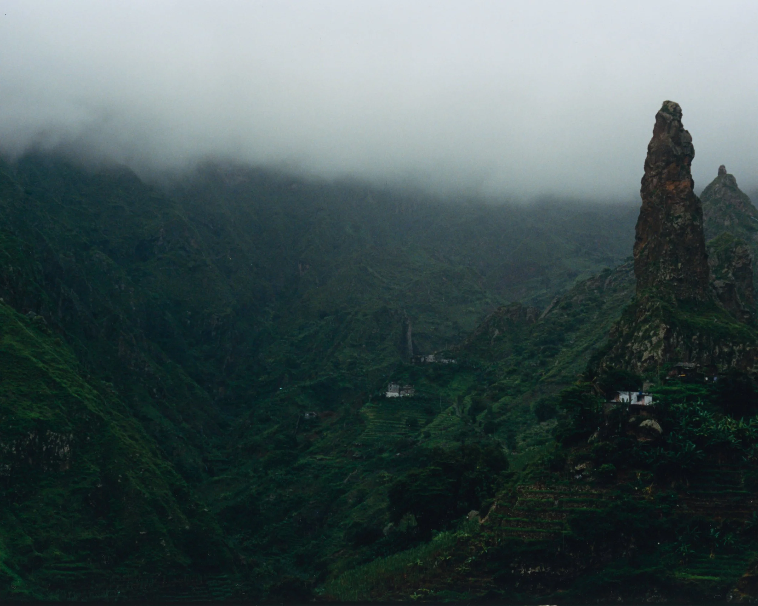 Paysage de montagnes verdoyantes, sommet rocheux imposant, brume couvrant la partie supérieure, petites maisons dispersées dans la vallée.