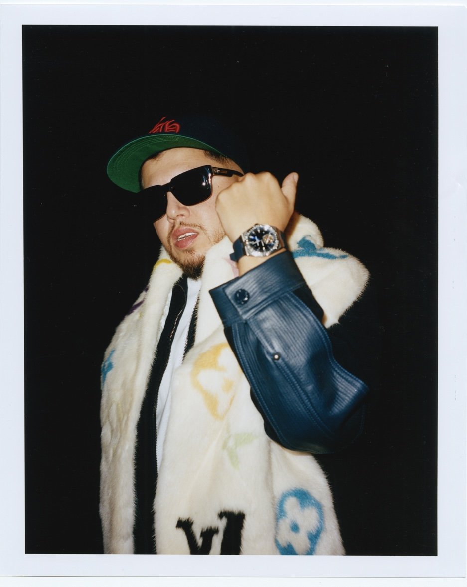 Homme portant des lunettes de soleil noires, une veste en fourrure faux, une casquette verte et un bracelet montre, posant avec une attitude confiante contre un fond noir.