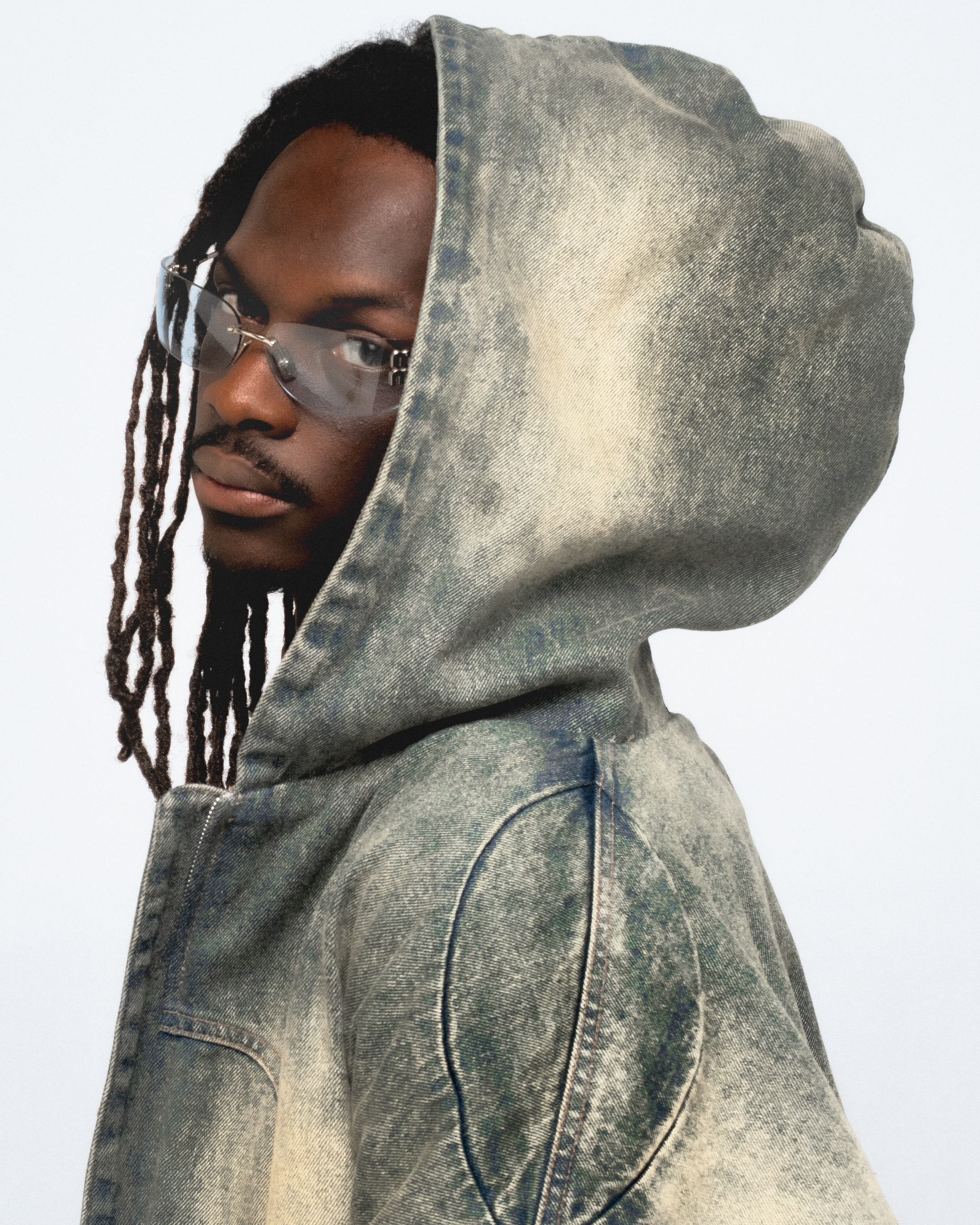 Homme portant une veste en jean délavée avec capuche, portant des lunettes de soleil, regardant vers la gauche, cheveux tressés.