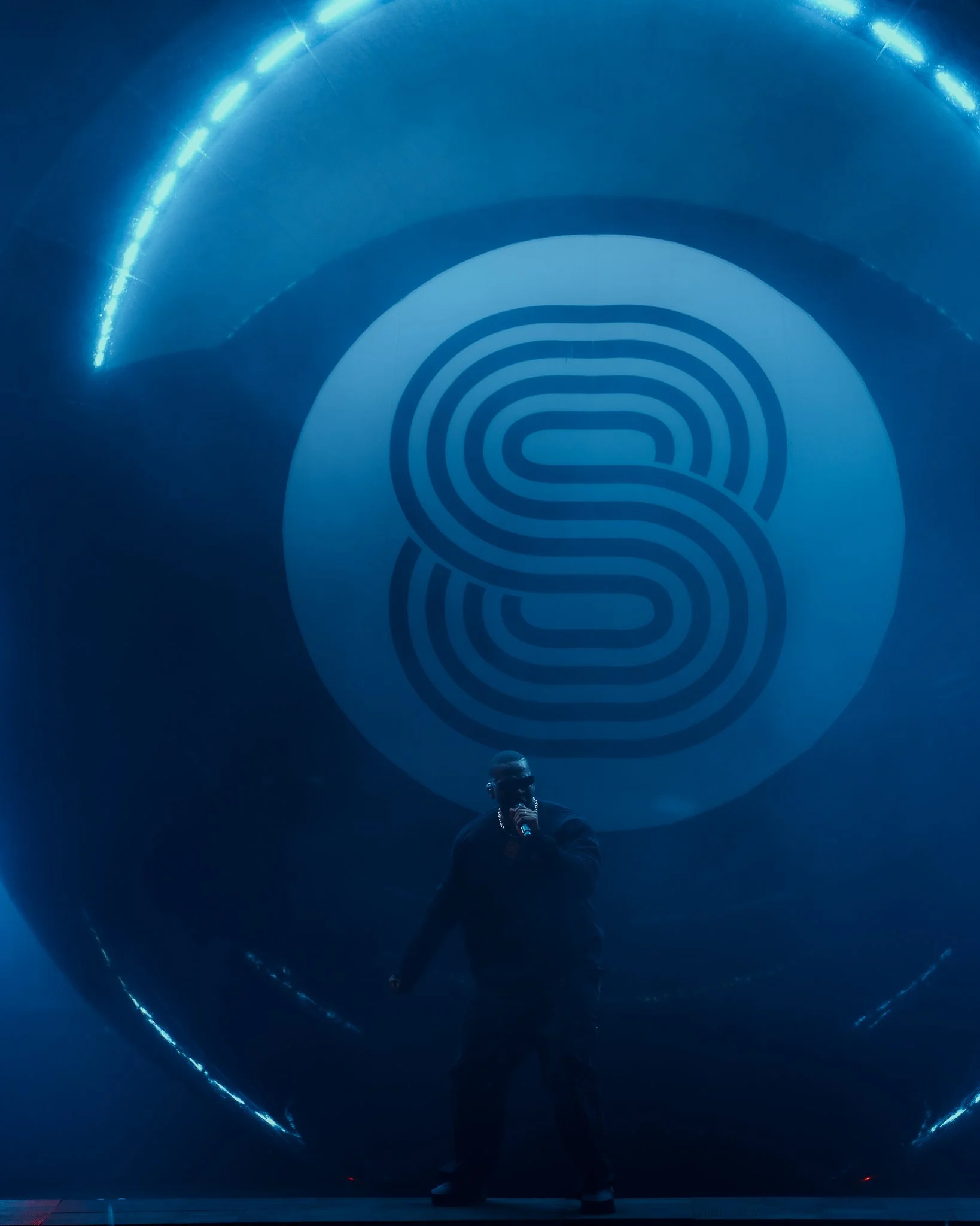 Un homme chante avec un microphone devant un grand logo lumineux de stylisé blanc sur fond bleu, dans un environnement sombre et moderne.