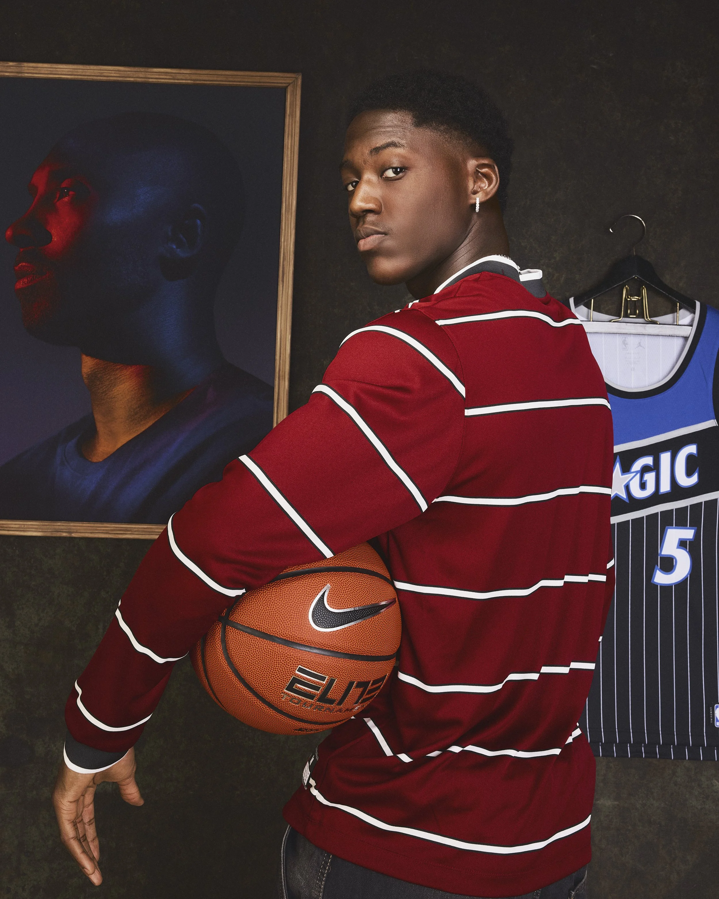 Un jeune homme pose avec un ballon de basketball Nike, portant un sweat rouge à rayures blanches, dans une pièce avec un portrait sur le mur et un maillot de basketball en arrière-plan.