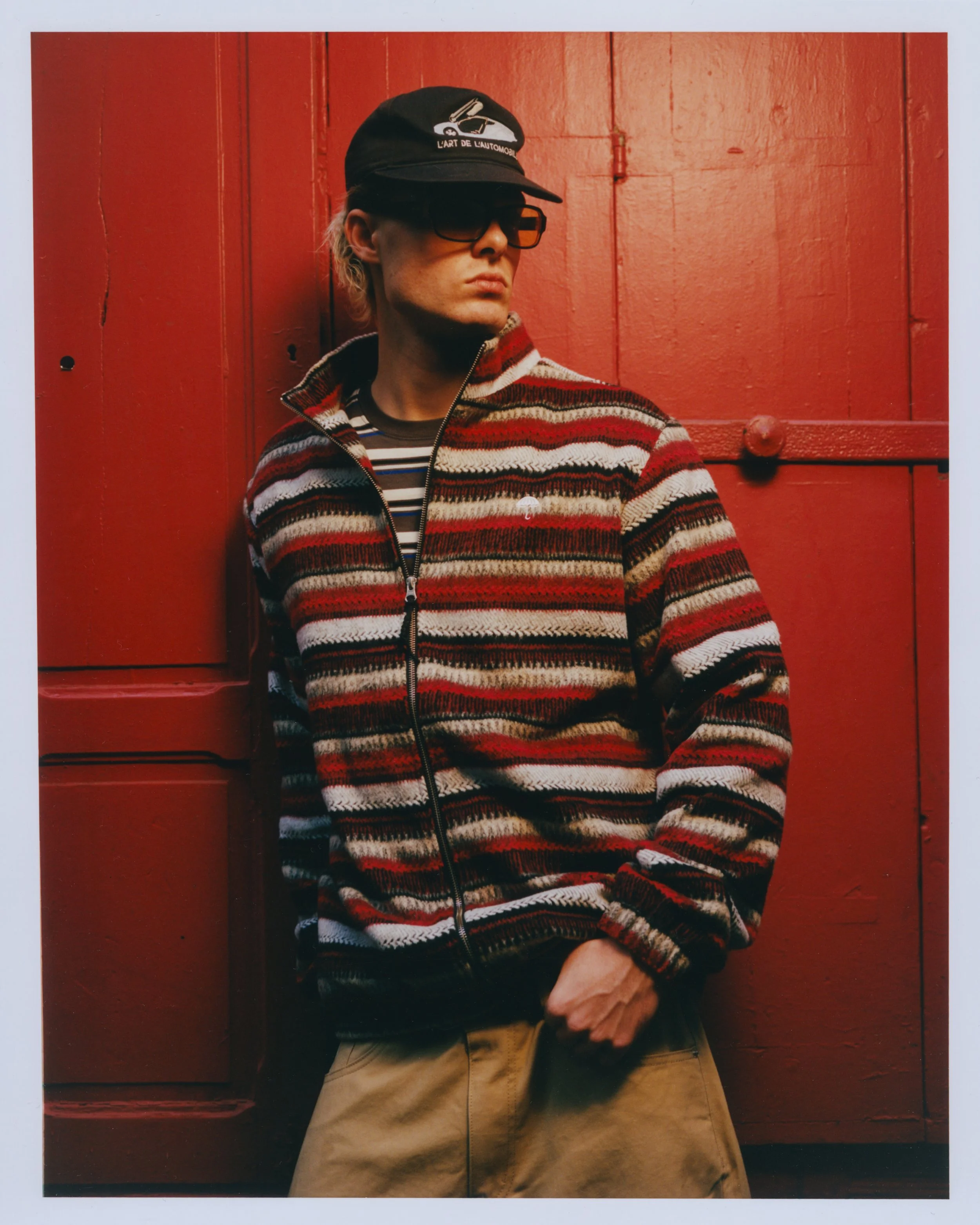 Un homme portant un casquette noire, des lunettes de soleil, un pull zippé à motifs rayés rouge, blanc, noir et beige, et un t-shirt rayé, posant contre un mur en bois rouge.